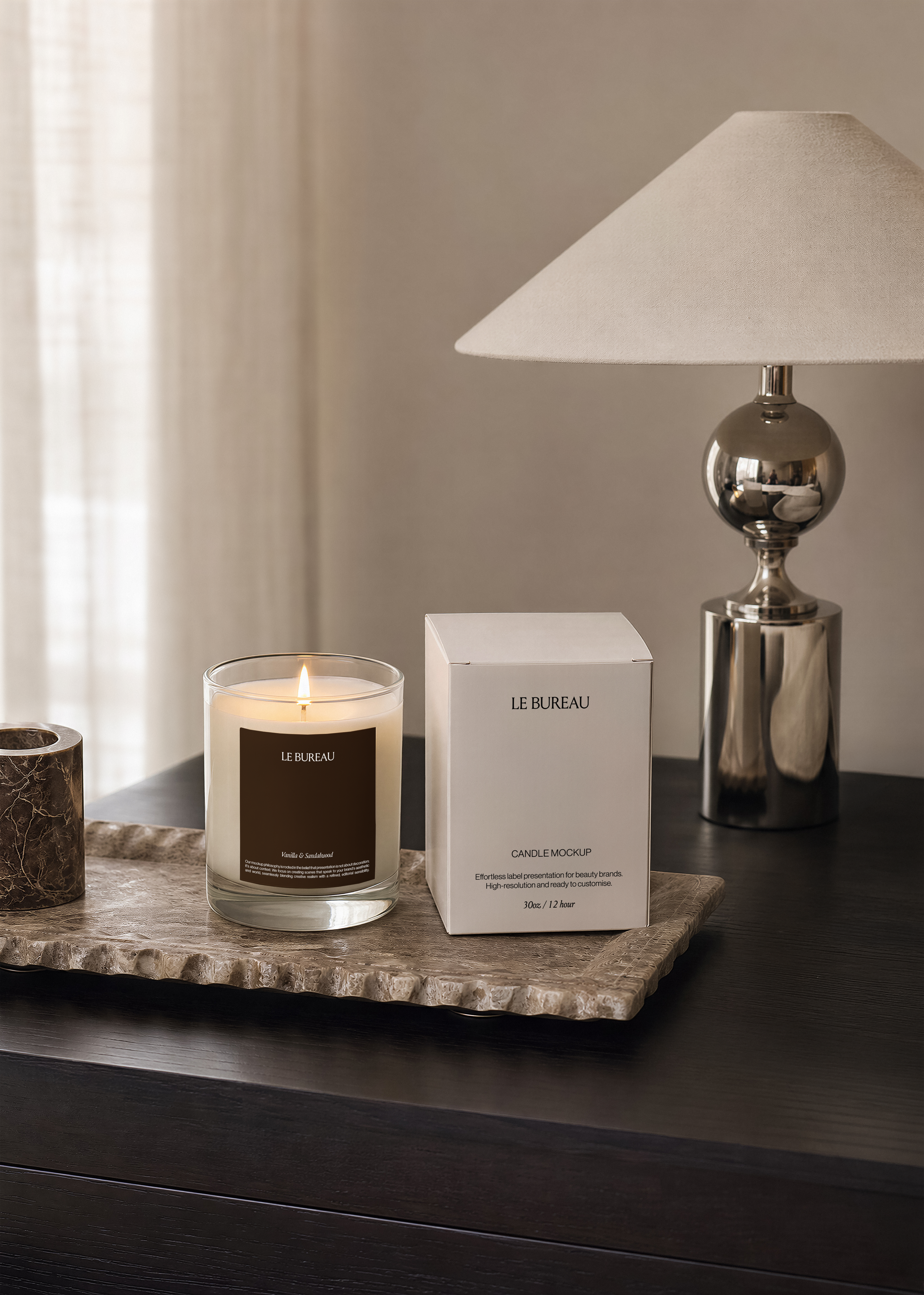 Lit glass candle jar mockup with pale label beside a silver table lamp, sheer curtain and warm neutral wall in soft focus