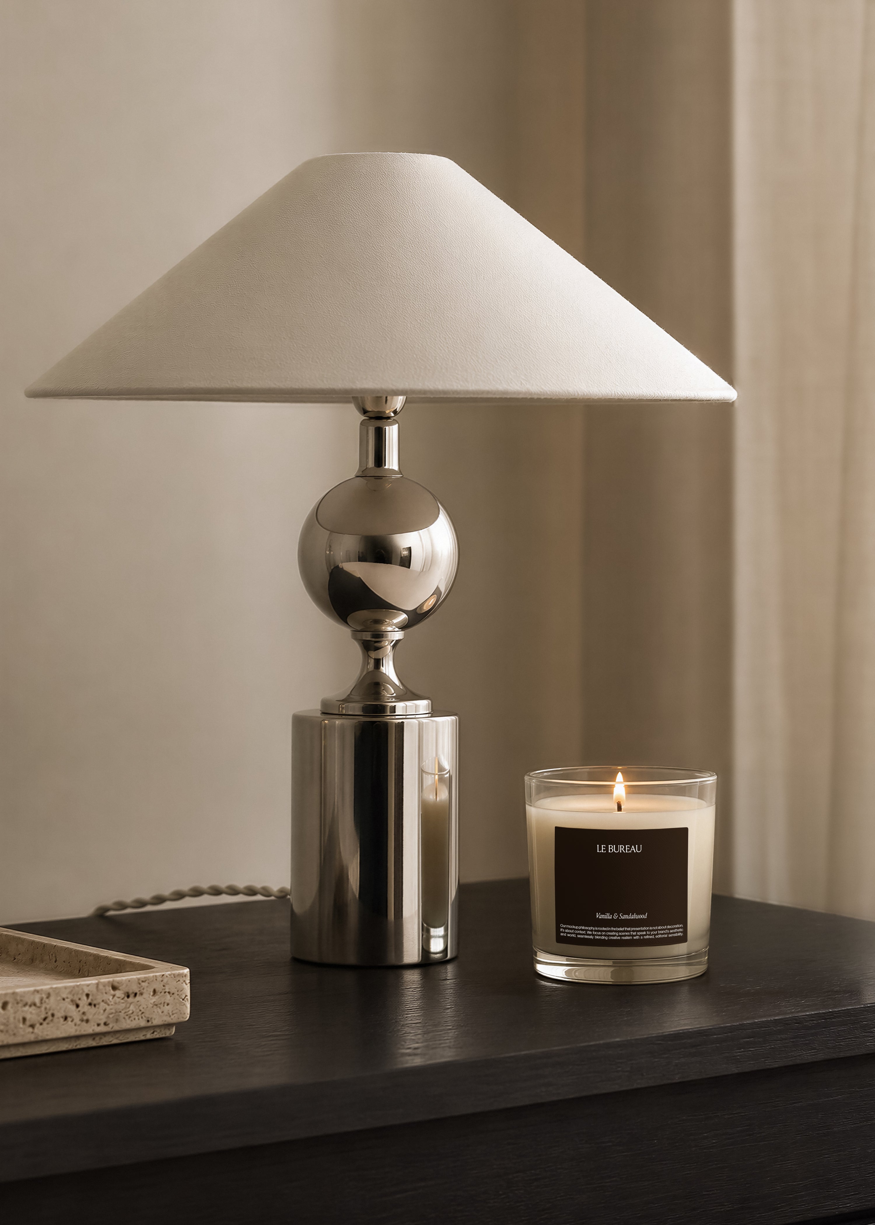 Glass candle jar mockup with dark brown label, positioned near a travertine tray and chrome lamp base