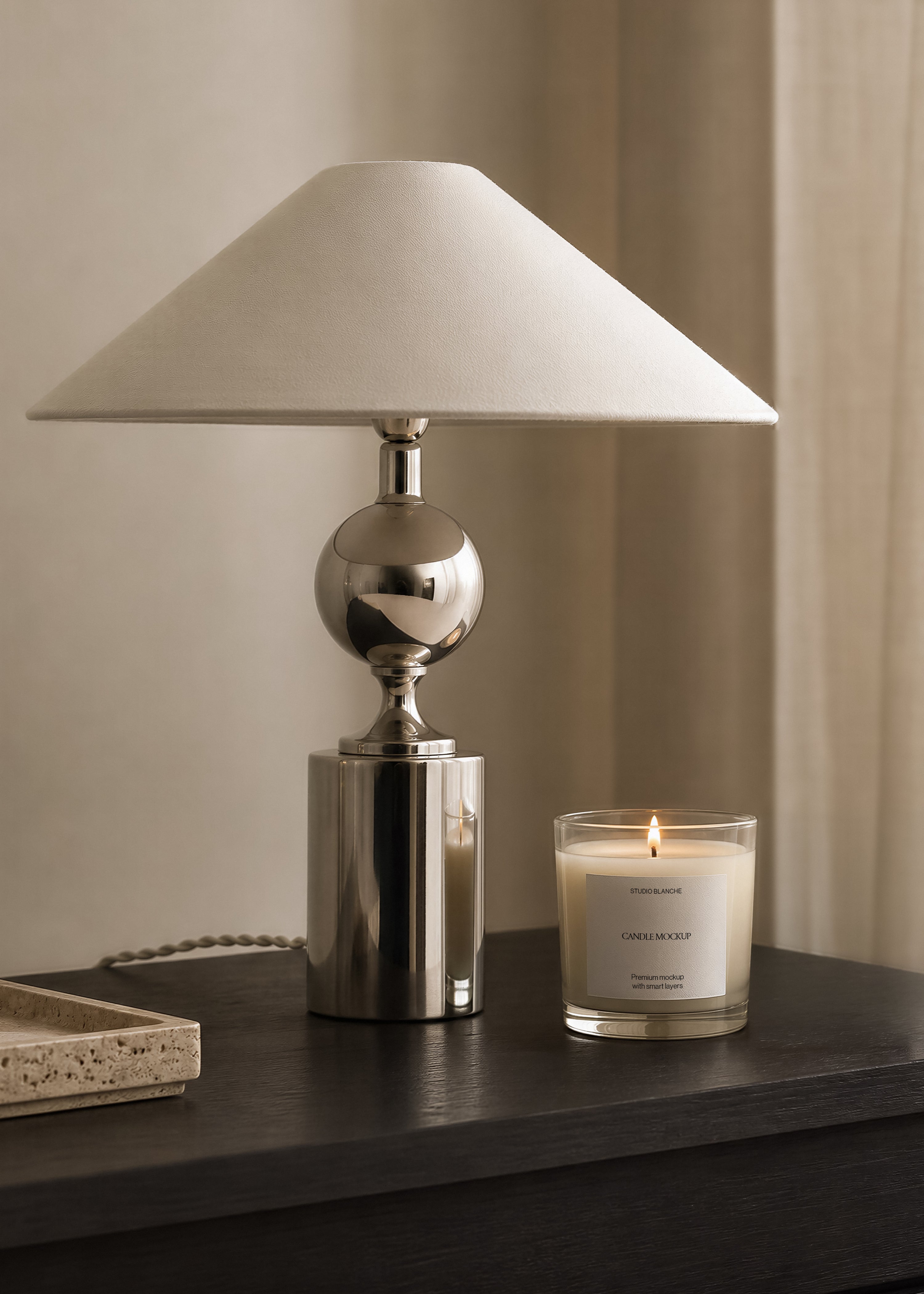 Lit glass candle jar mockup beside a sculptural chrome table lamp on dark wood surface, warm neutral curtain in background