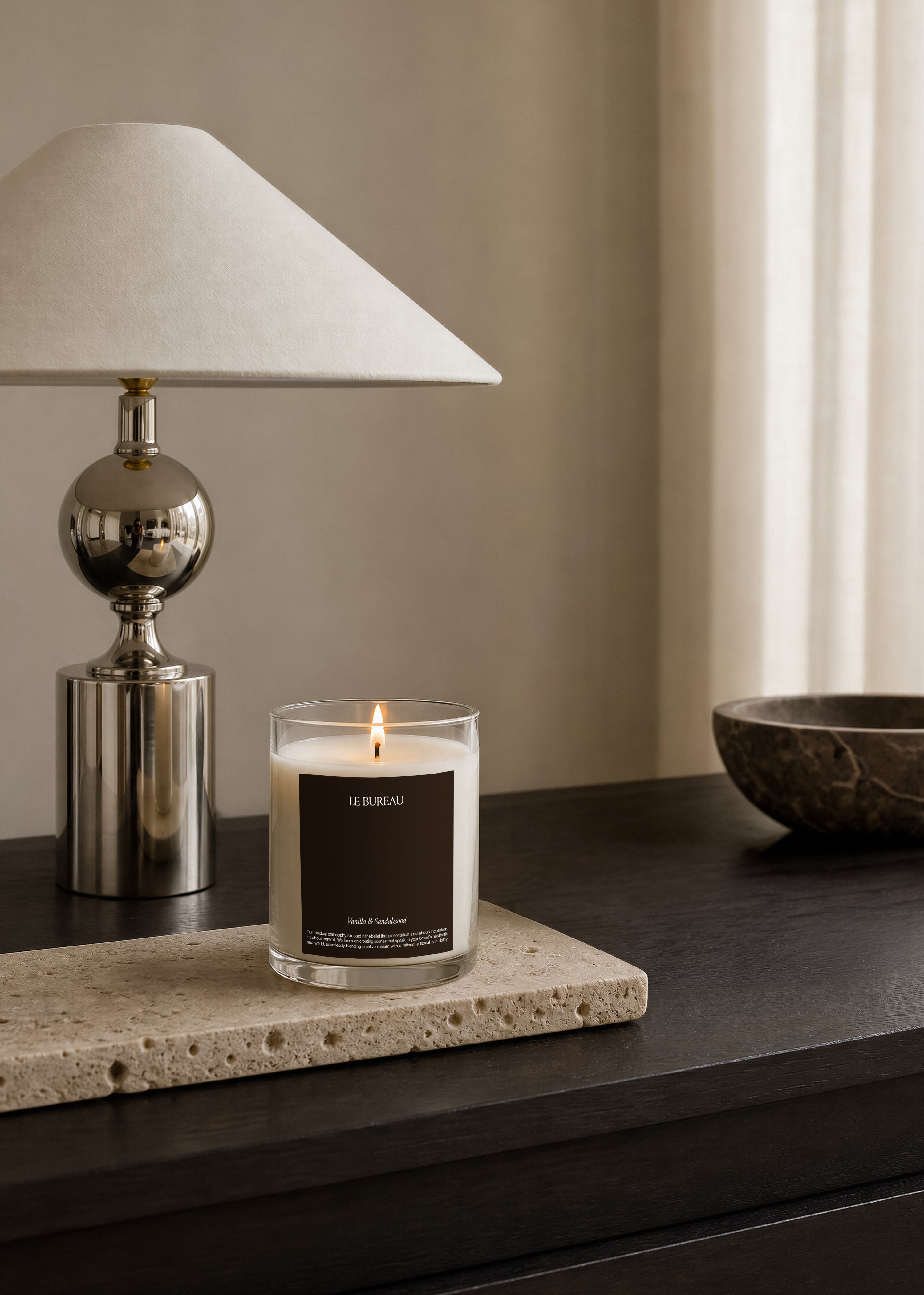 Lit glass candle jar mockup with dark label on a travertine slab, chrome table lamp and dark marble bowl in warm interior setting