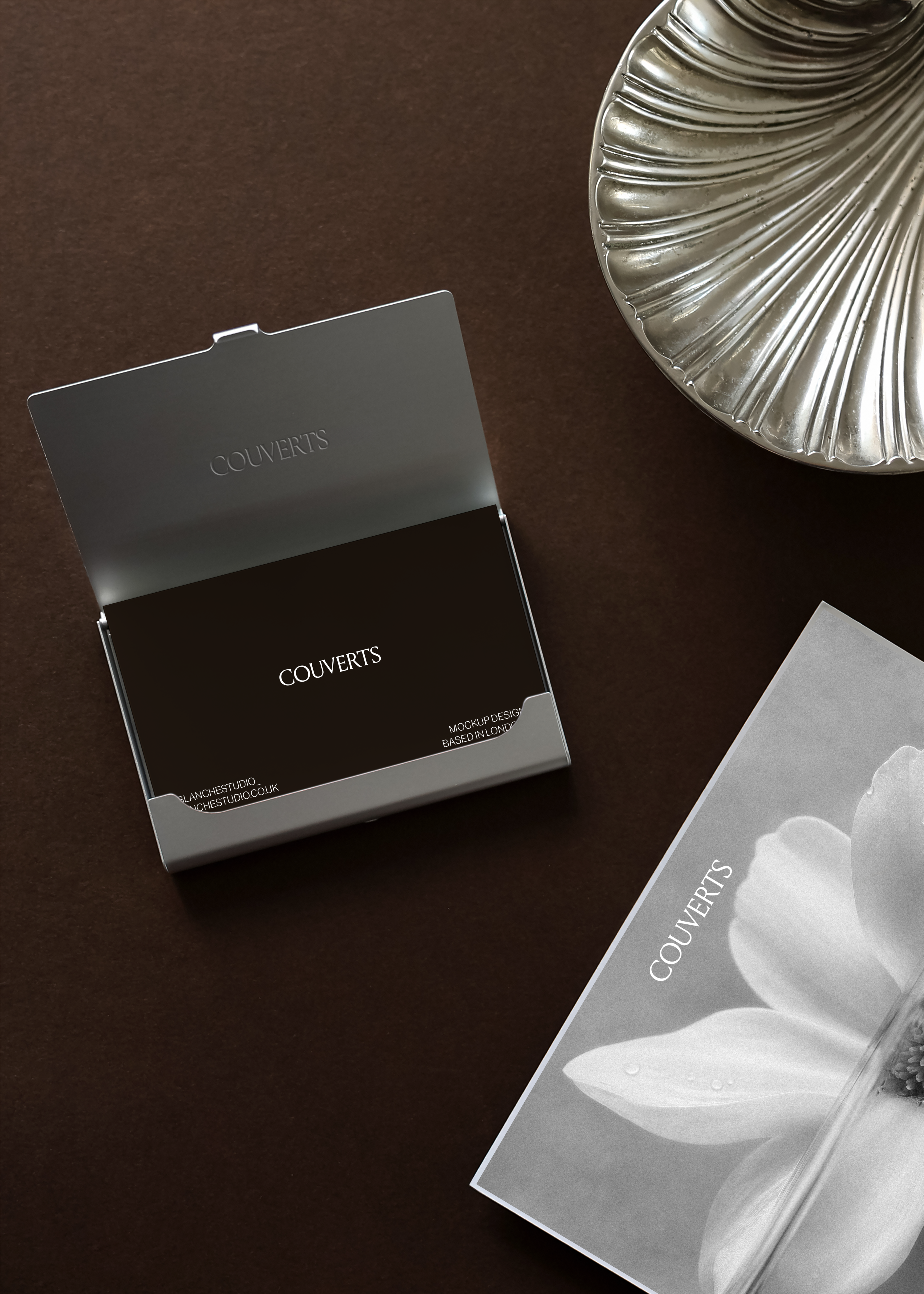 Business card mockup in open metal card holder beside ornate silver shell dish and A6 card, dark editorial flat lay