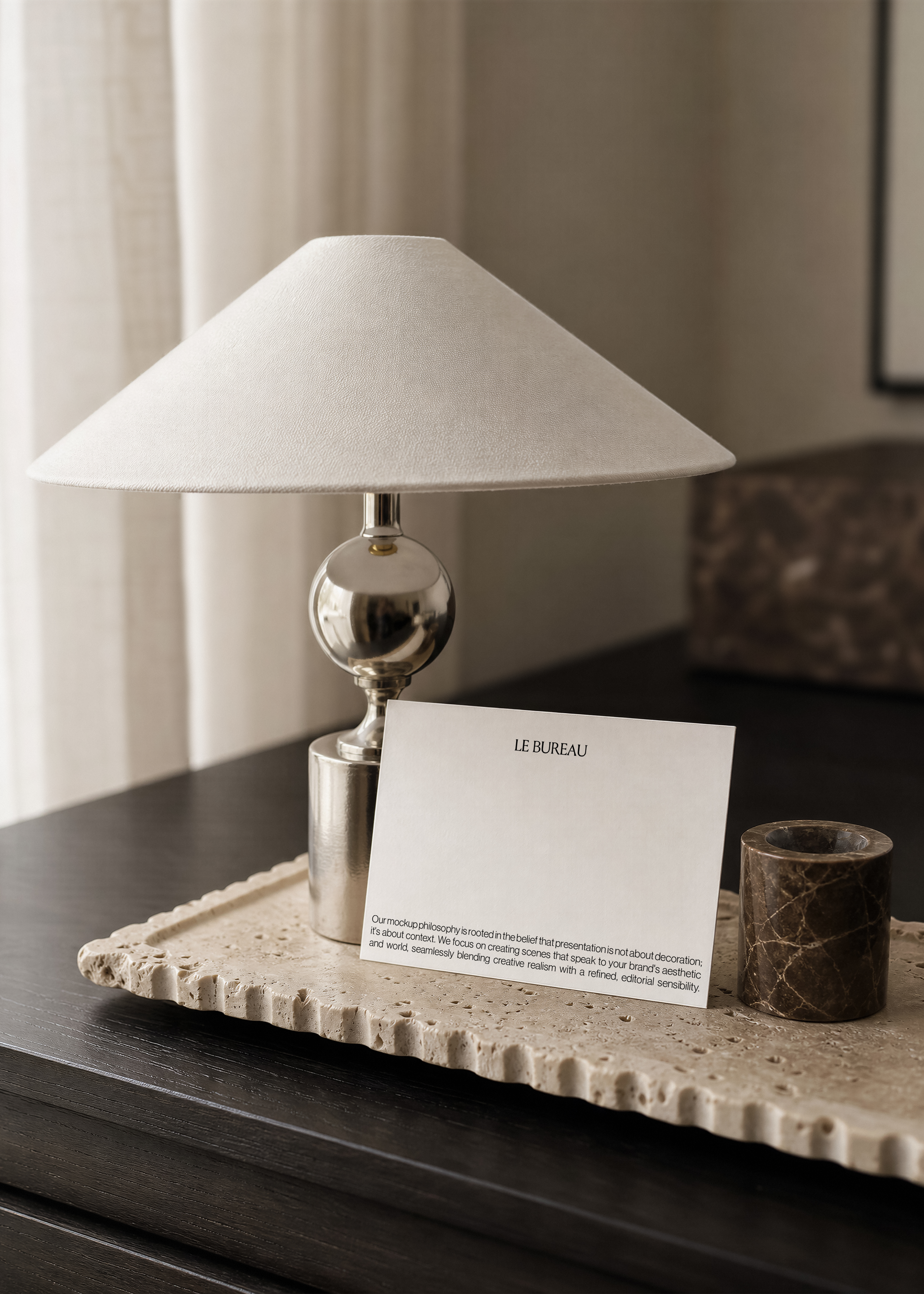 Card mockup with minimal wordmark propped against a chrome table lamp on a travertine tray, dark marble candle holder alongside