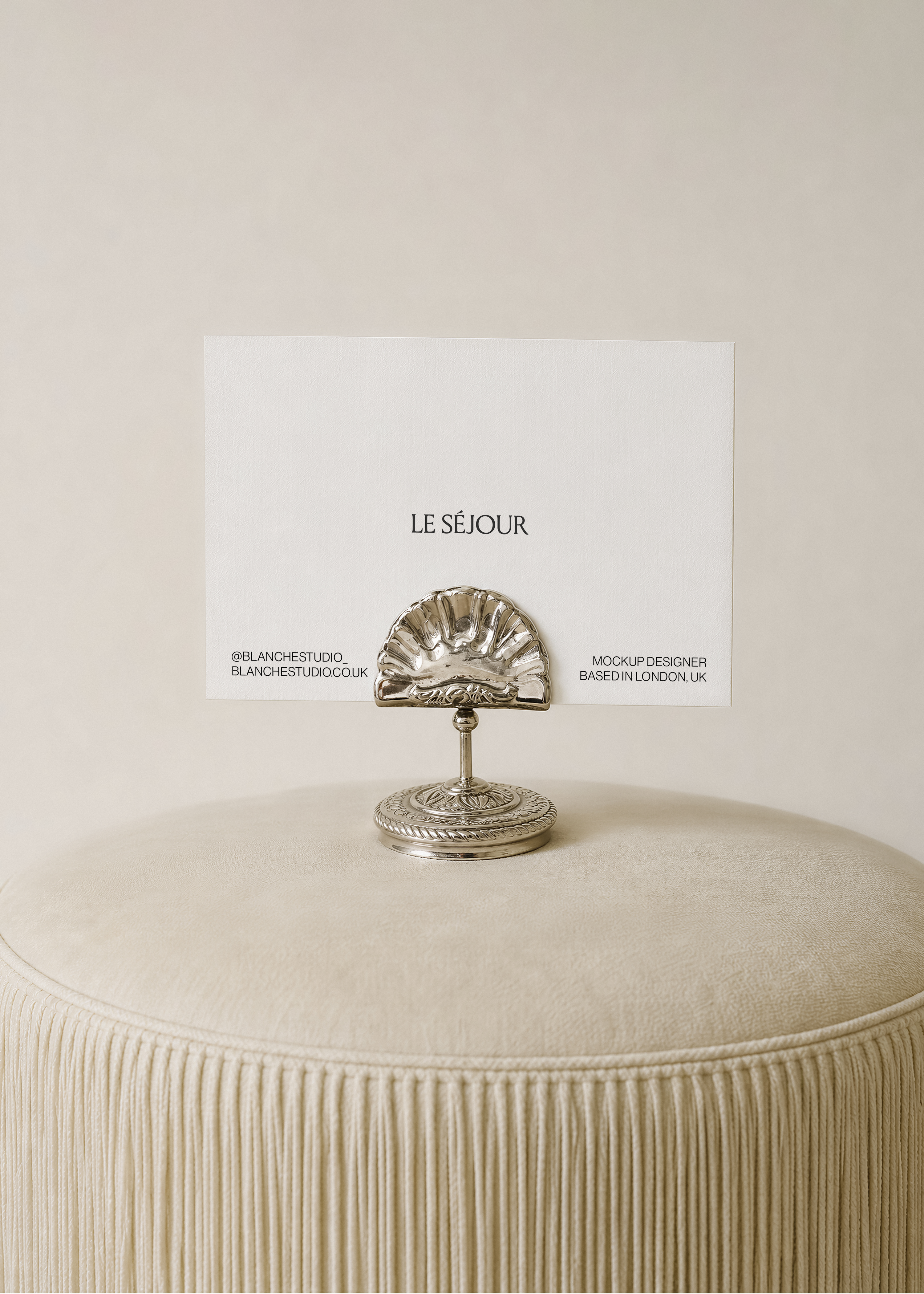 Landscape card mockup held upright in an ornate silver shell card holder on a cream fringed pouf against a neutral wall