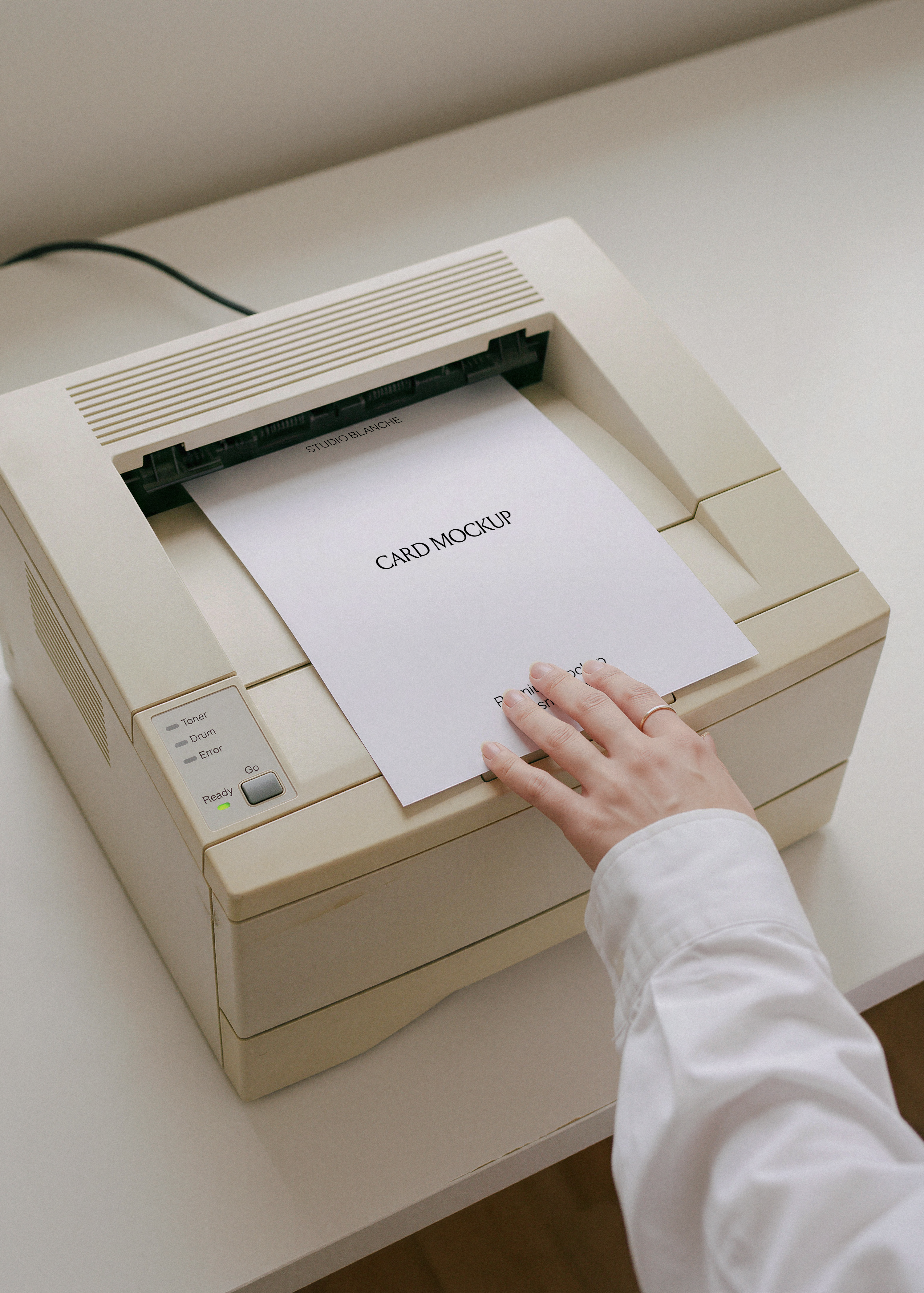 Blank A4 card printing from retro beige laser printer with Ready light on, hand resting on paper, pale desk surface
