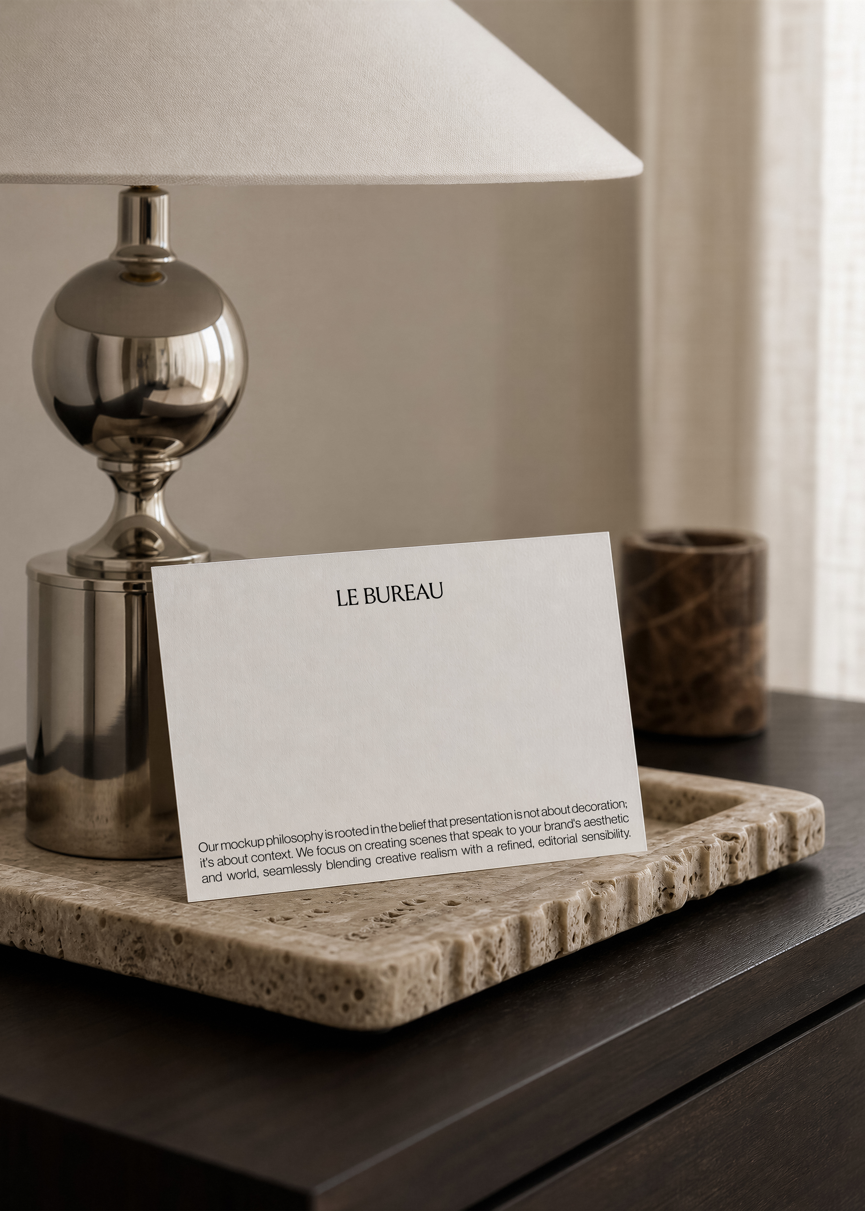 Card mockup with printed wordmark leaning against a chrome lamp base on a textured travertine tray, dark wood surface below