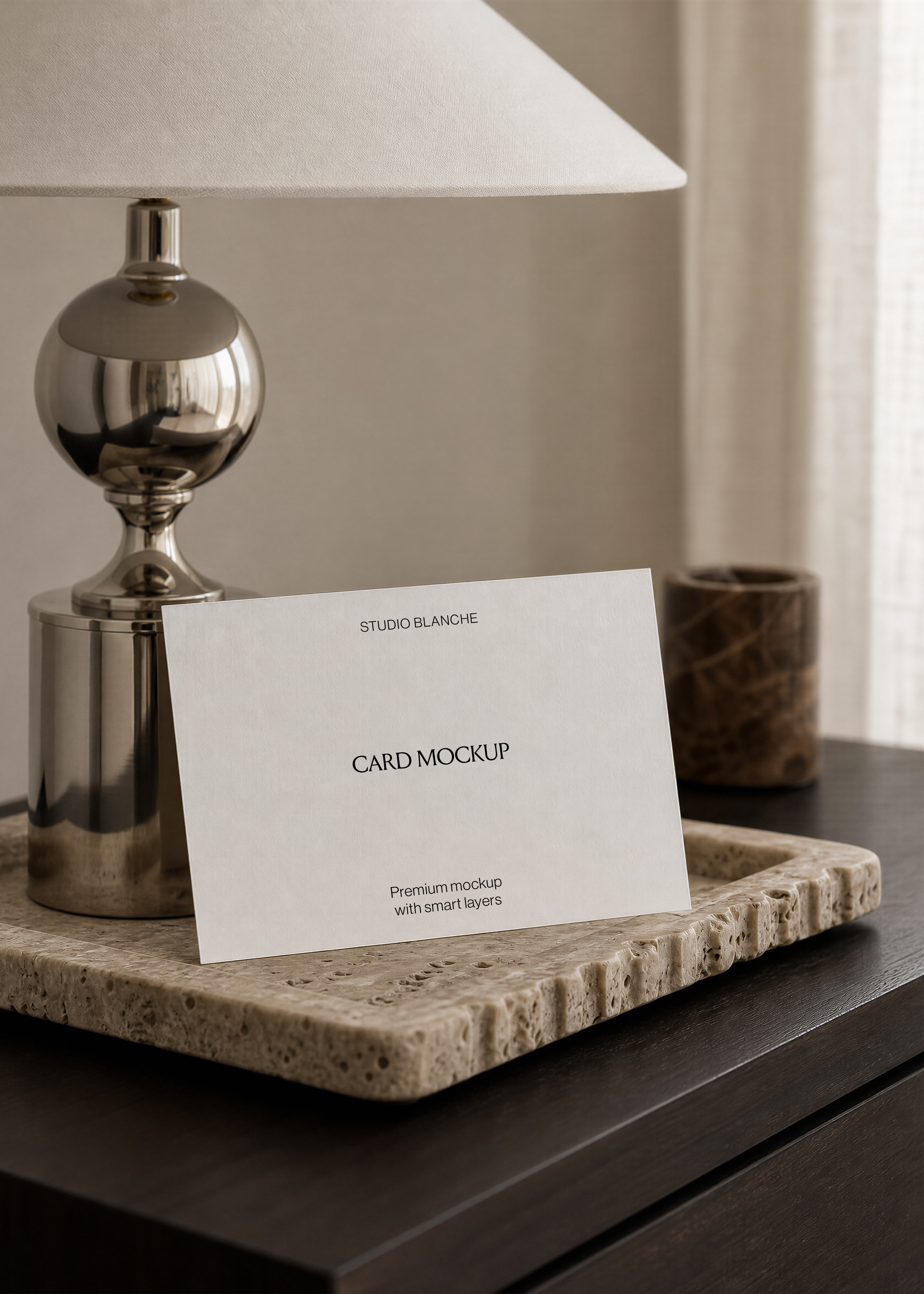 Blank card mockup with small printed text, propped upright beside a marble candle holder on travertine