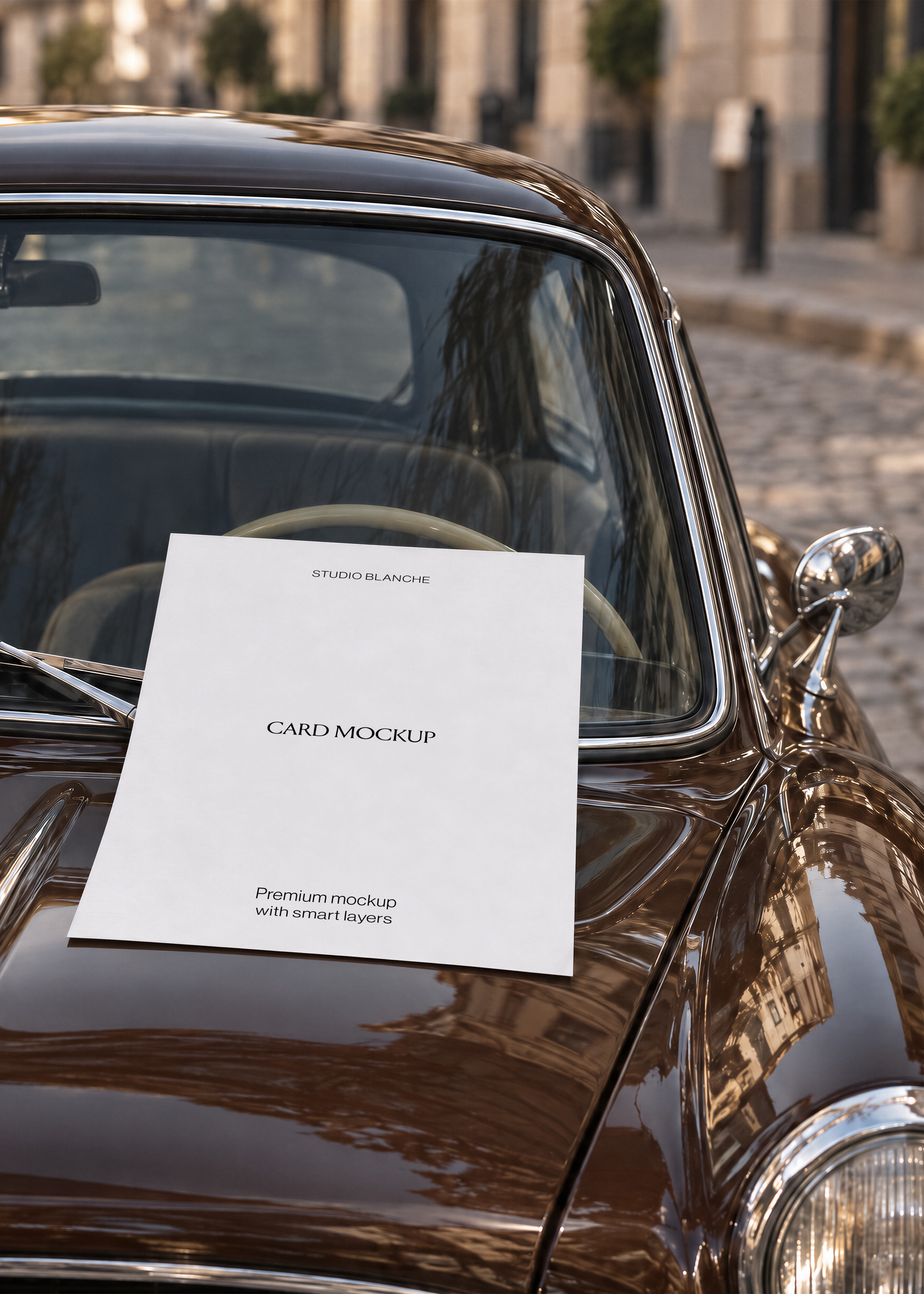 Blank white card mockup resting on glossy brown vintage car bonnet against windscreen, chrome mirror, cobbled street