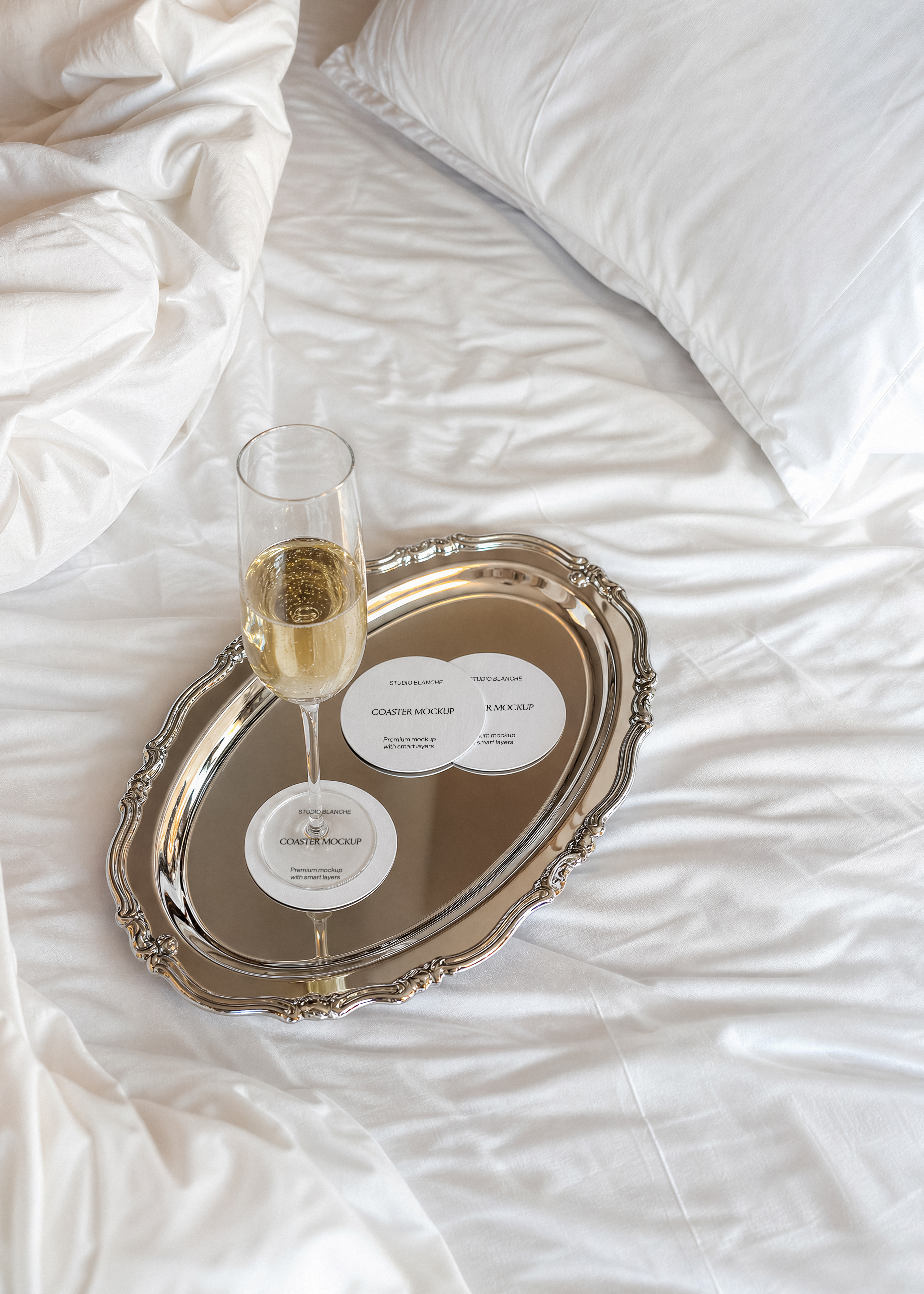 Coaster mockup trio arranged on a mirrored silver tray, champagne glass resting on one, creased white sheets