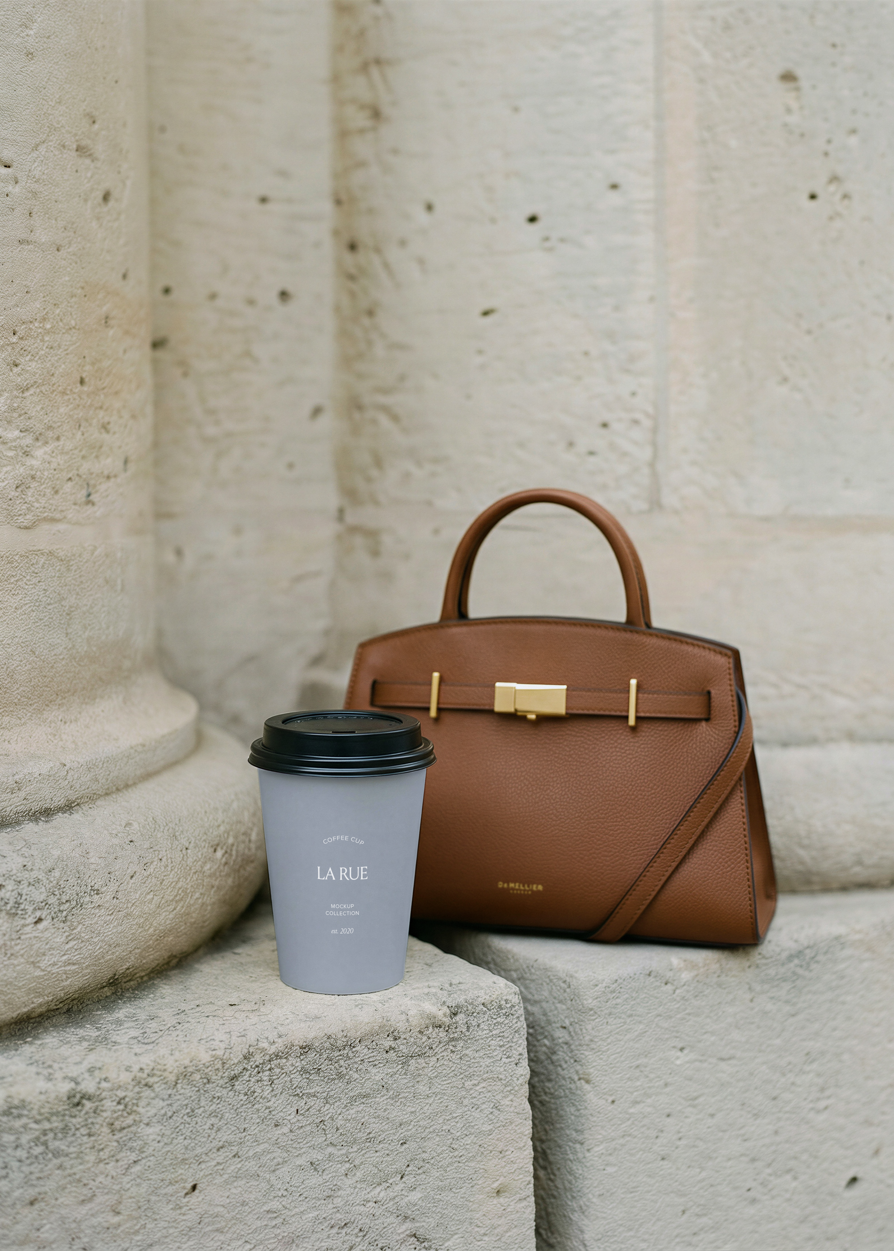 Takeaway coffee cup with grey branded sleeve and serif logo on stone steps beside a tan leather handbag, from Studio Blanche