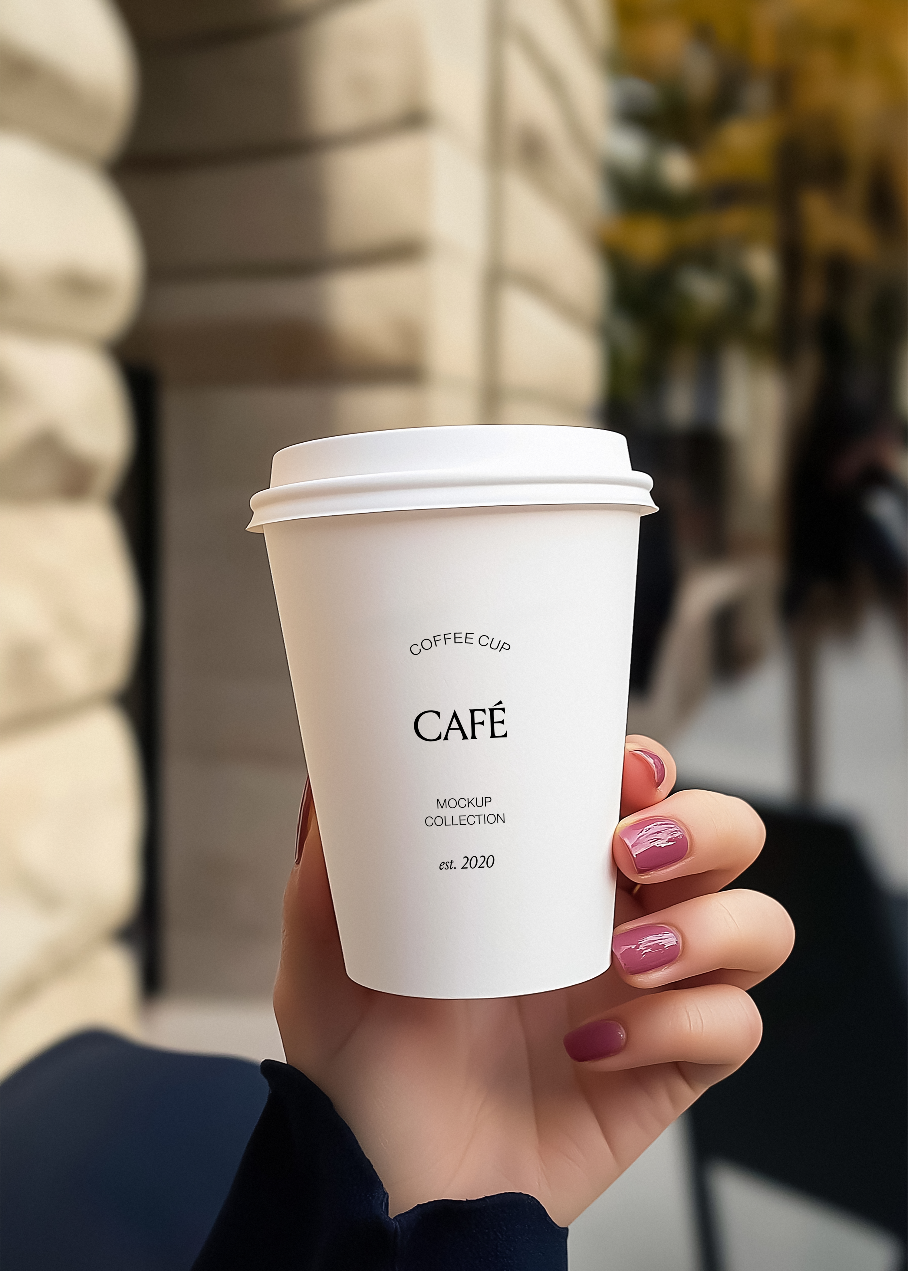 Hand holding a white takeaway coffee cup mockup on a sunlit street, blurred stone building and café seating behind