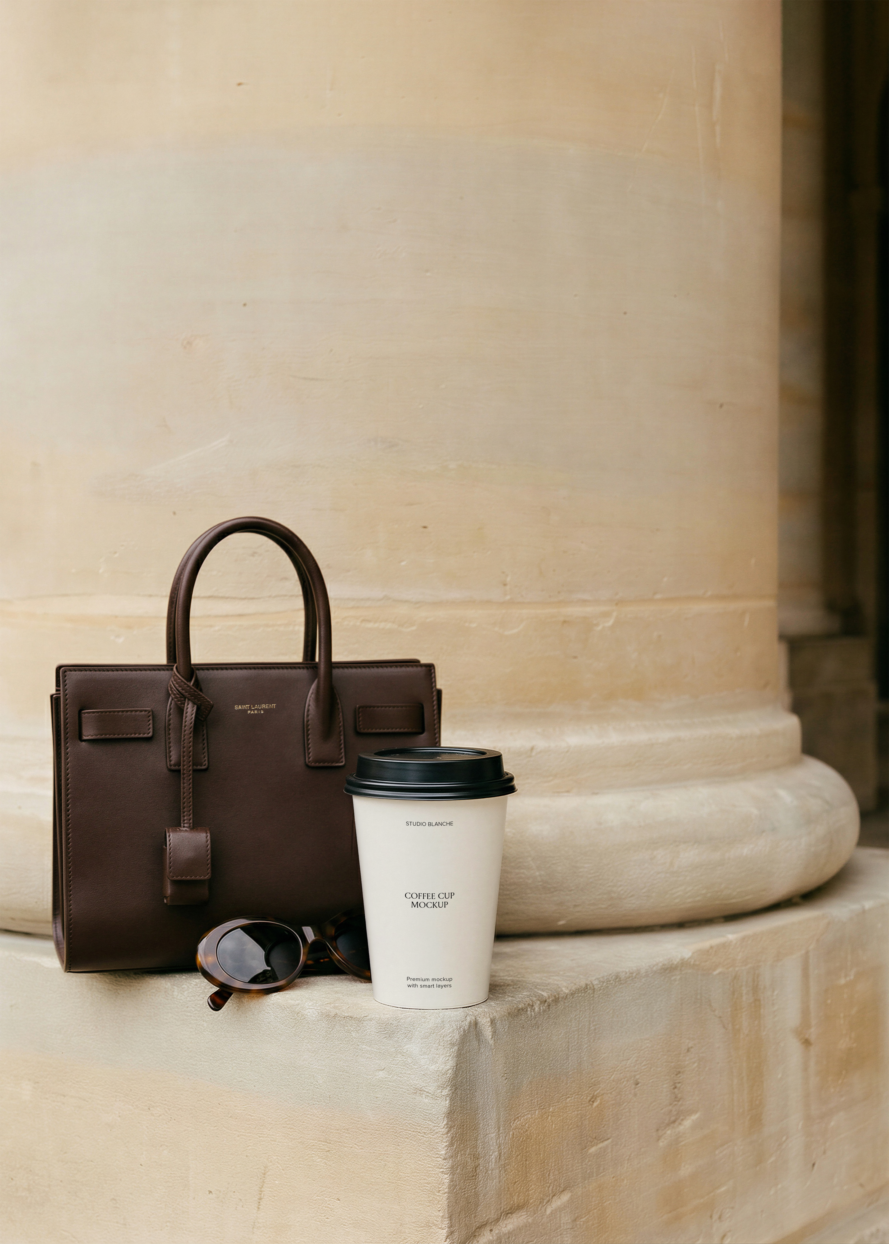 Blank white takeaway coffee cup with black lid on a stone column base beside a dark brown leather handbag and sunglasses, from Studio Blanche