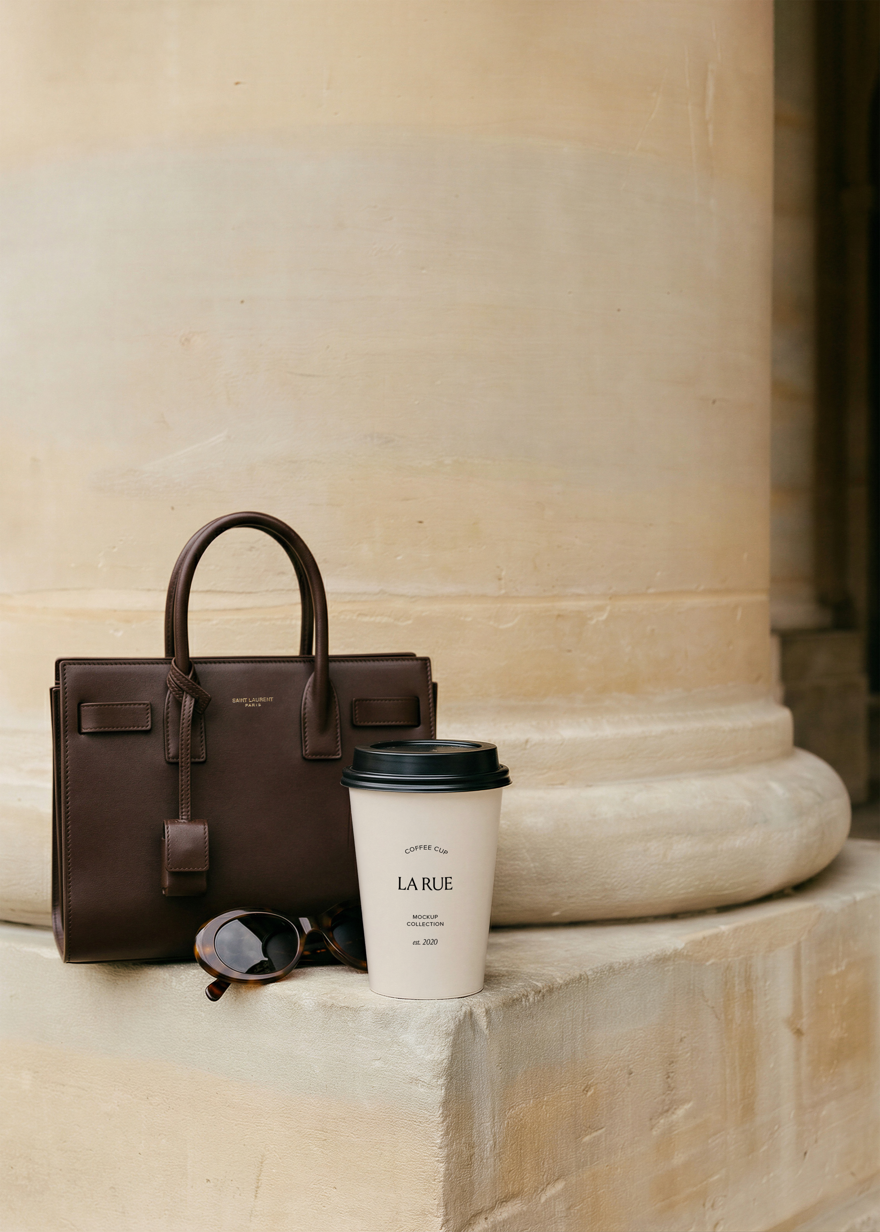Takeaway coffee cup with serif brand name on stone column base beside a dark brown leather handbag and tortoiseshell sunglasses, from Studio Blanche