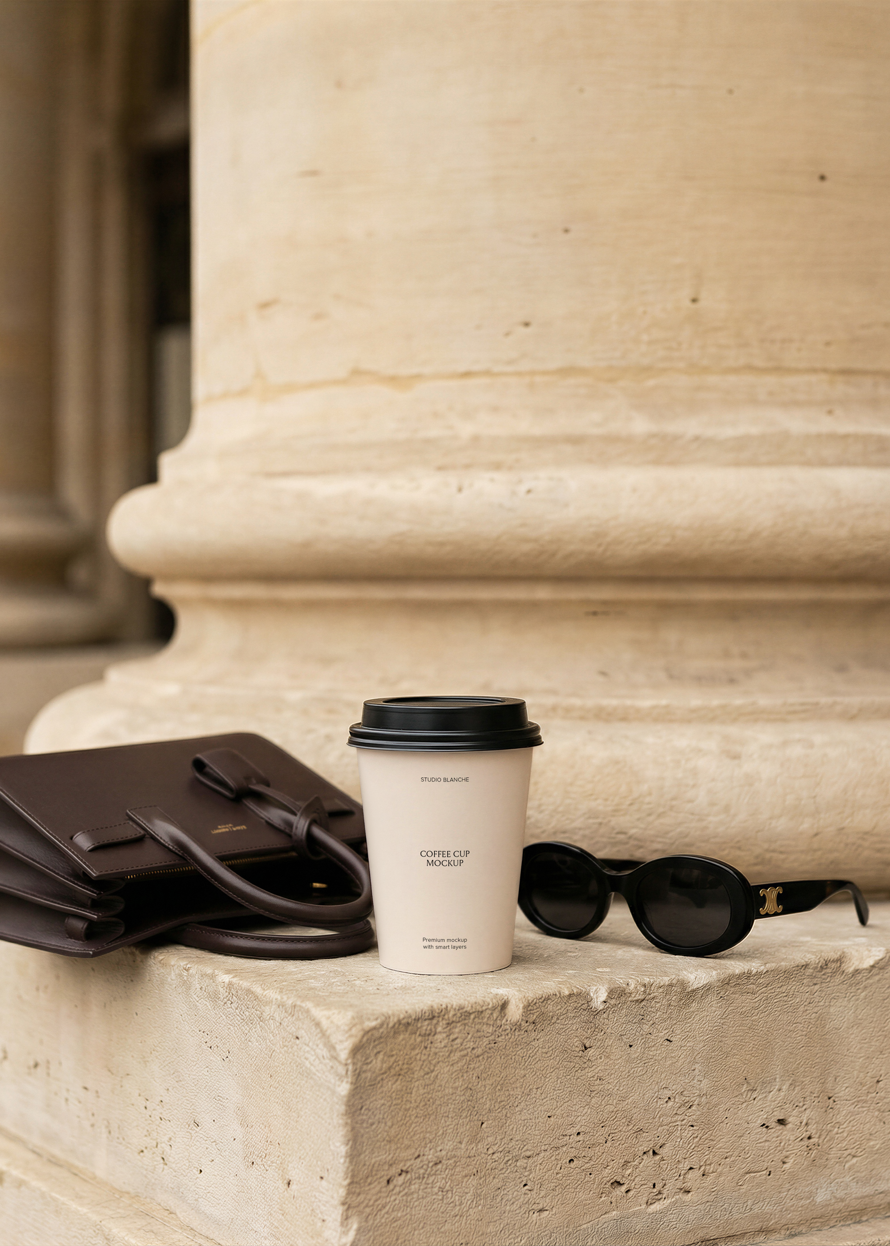 Blank white takeaway coffee cup with black lid on stone steps beside a dark leather bag and black sunglasses against a limestone column, from Studio Blanche