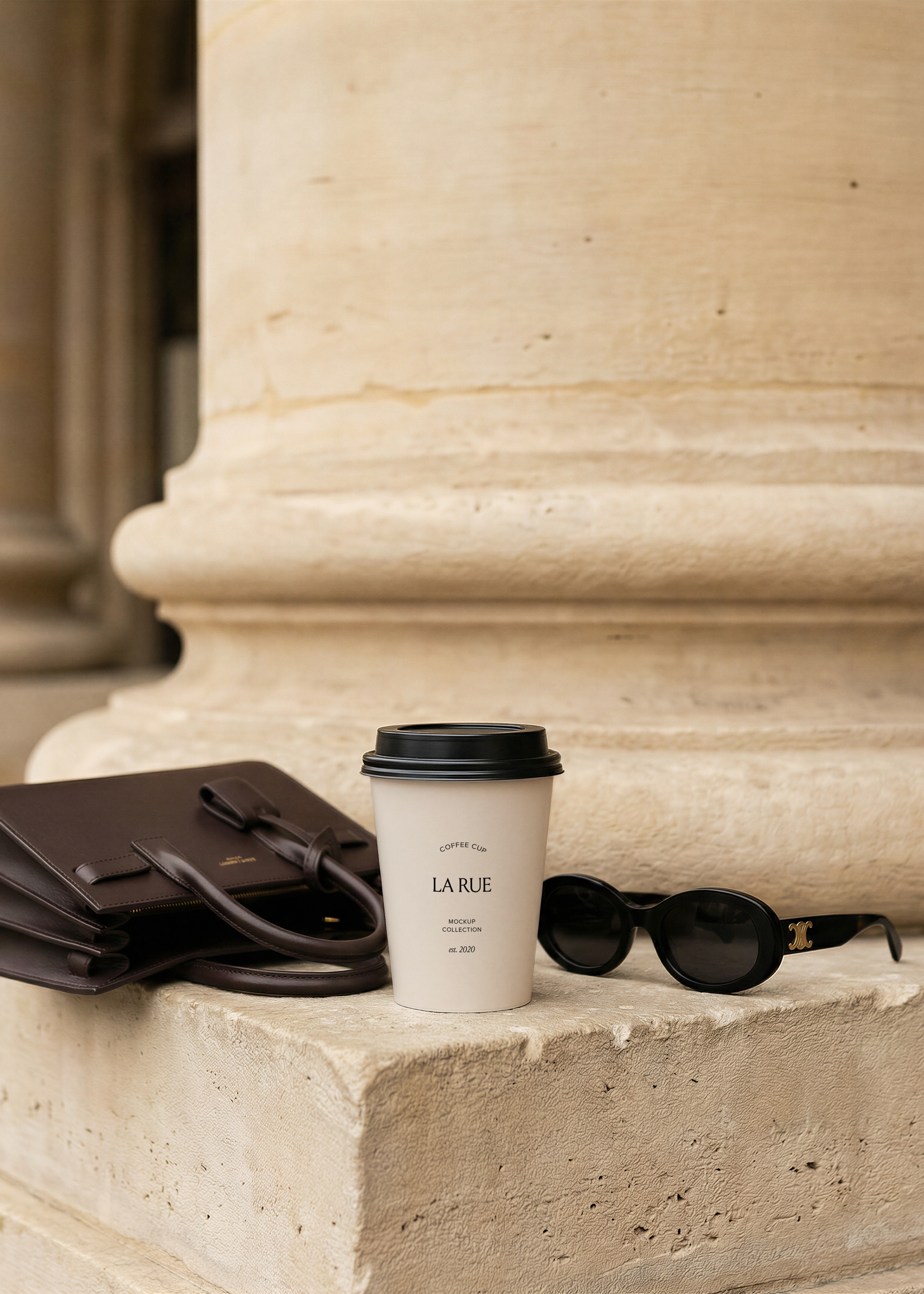 Takeaway coffee cup with serif brand name on stone steps beside a dark leather bag and black oval sunglasses against a limestone column, from Studio Blanche