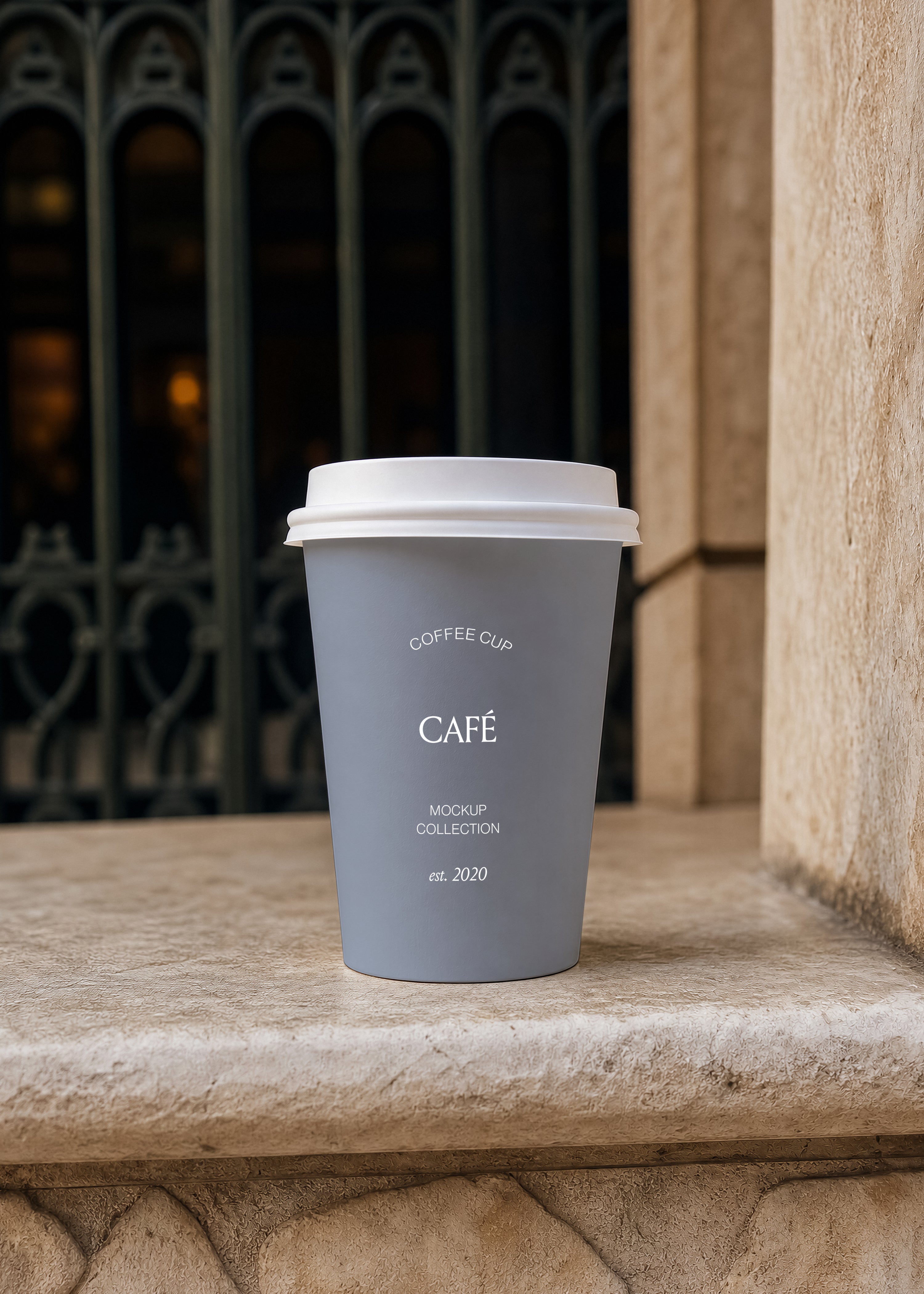 Matte blue takeaway coffee cup with white lid on weathered stone ledge, dark ironwork gate and warm interior light behind