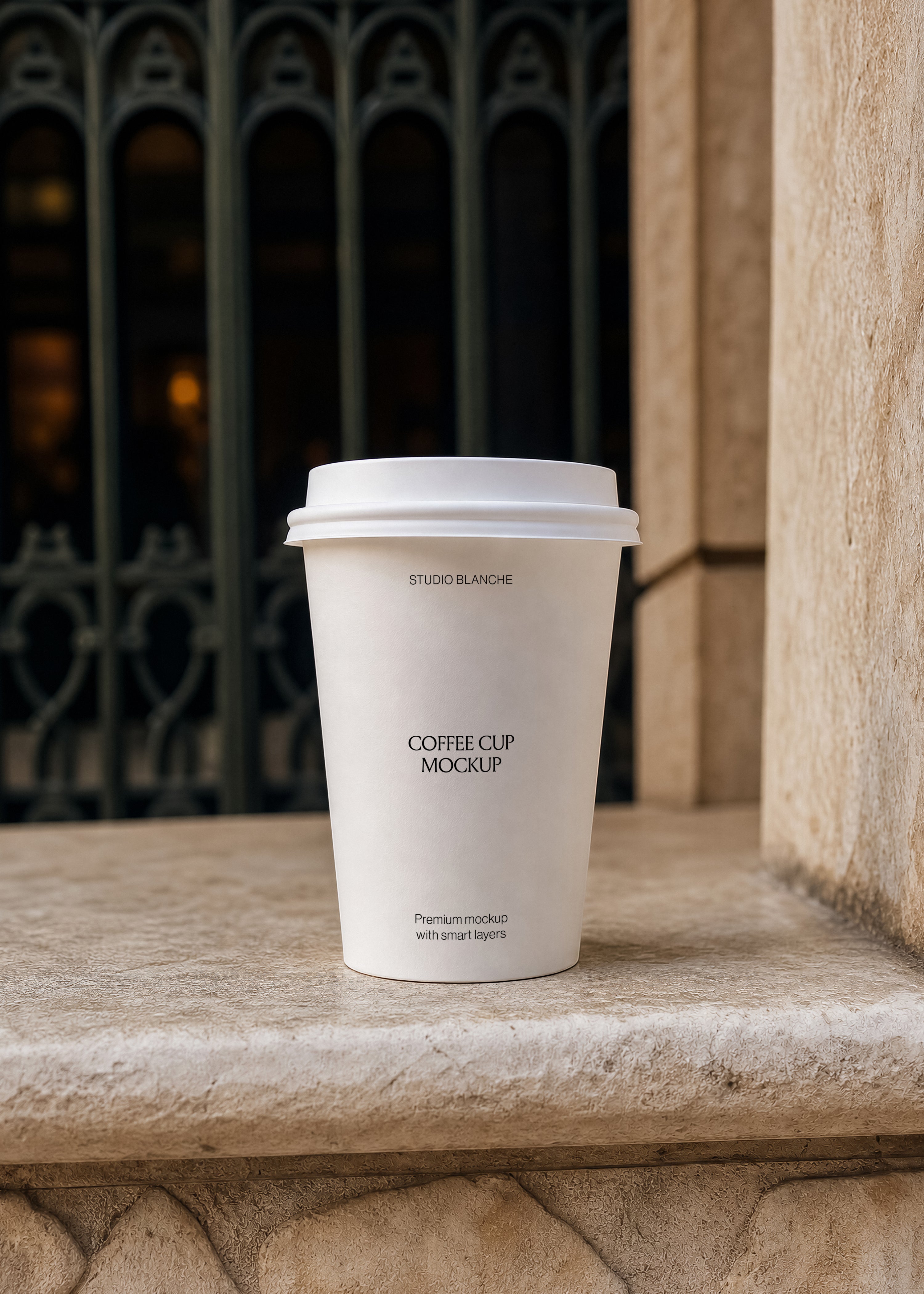 Cream takeaway coffee cup mockup with STUDIO BLANCHE branding on stone ledge, same ironwork doorway backdrop