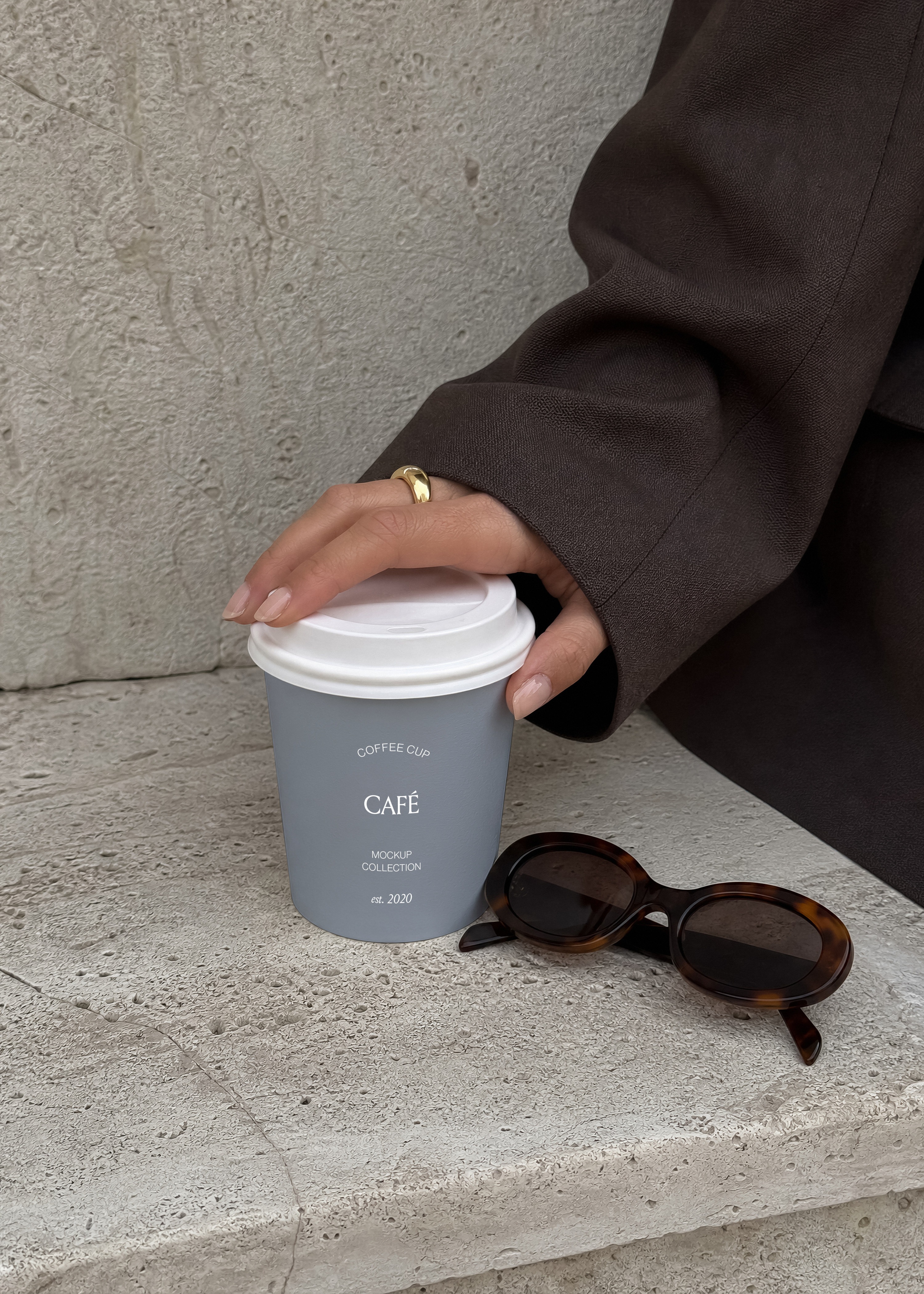 Coffee cup mockup with white lid held by a hand against raw concrete, tortoiseshell sunglasses beside it, editorial styling