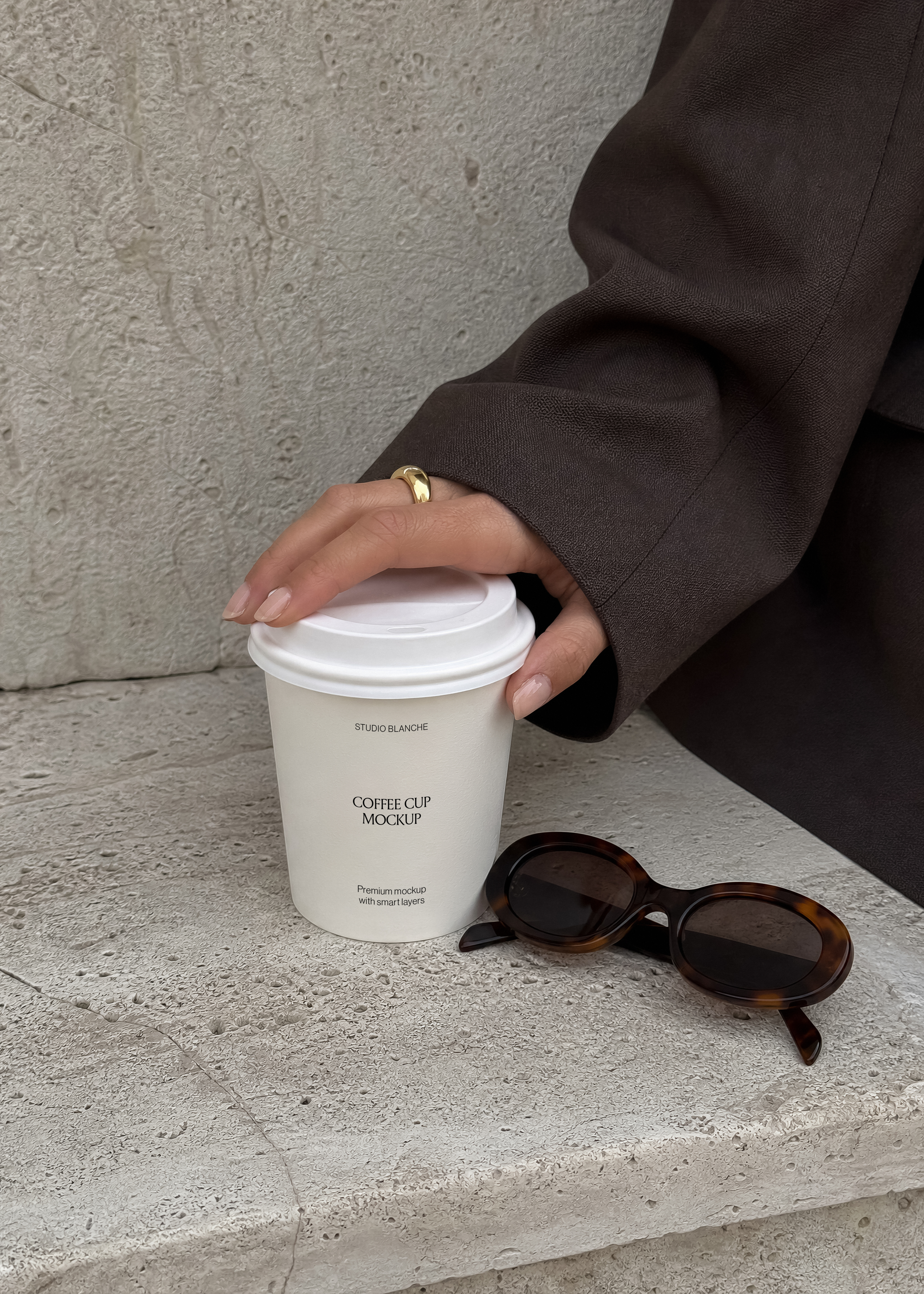 Close-up coffee cup mockup label area held mid-reach on concrete step, sunglasses and coat sleeve in frame