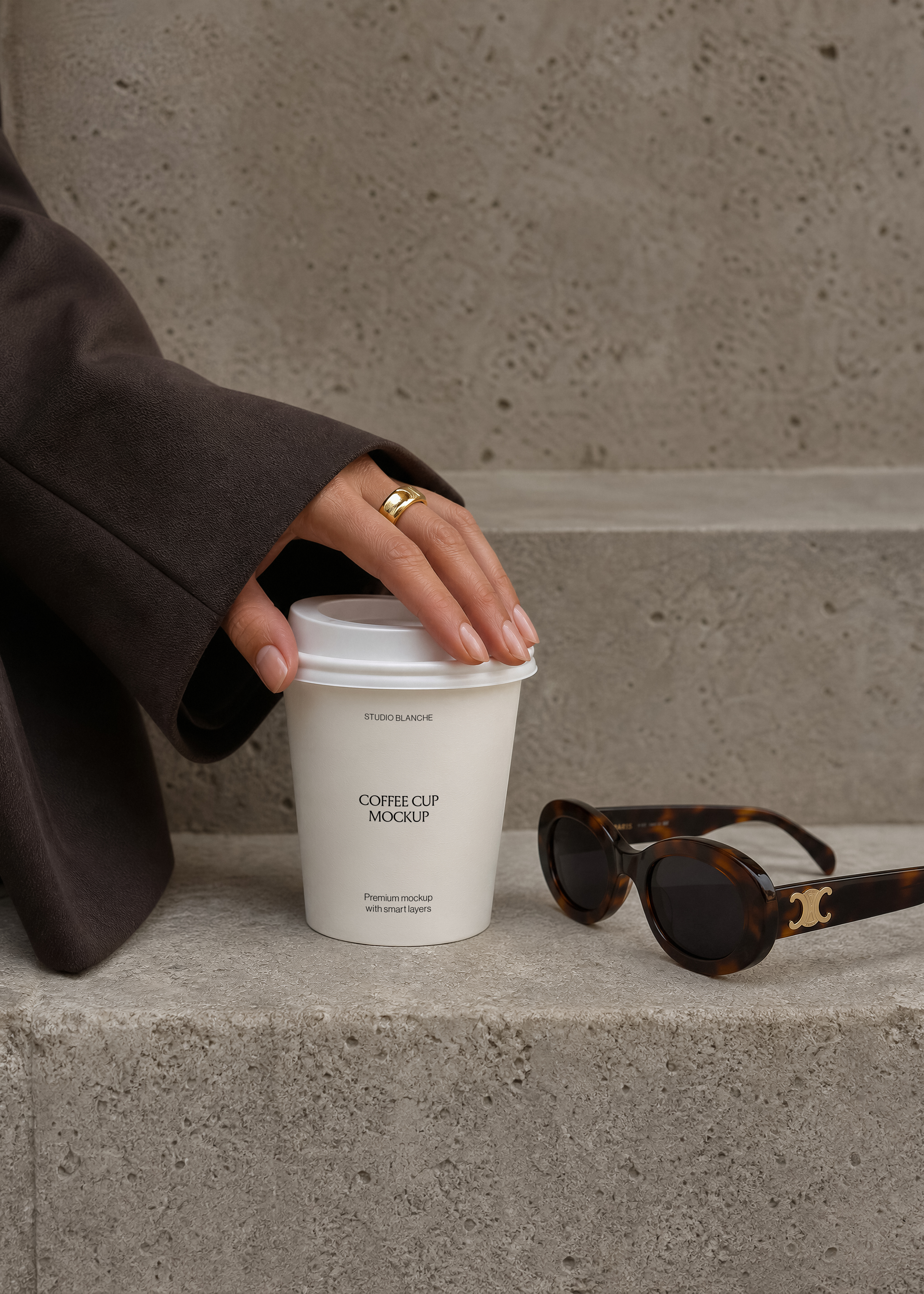 Close-up coffee cup mockup label on concrete step, hand with gold ring, dark coat sleeve, sunglasses with metal logo detail