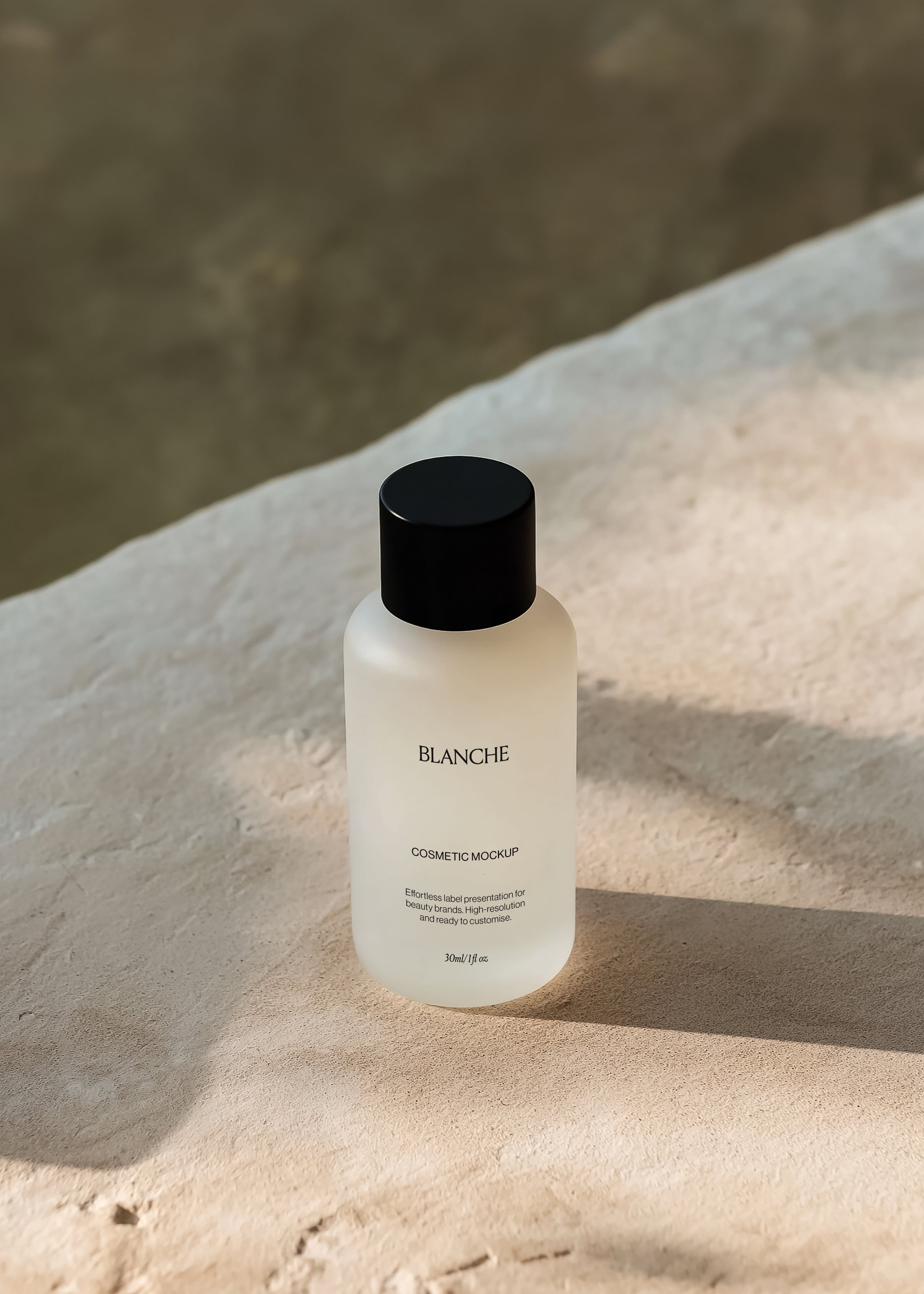 Frosted glass cosmetic bottle mockup with matte black cap on pale stone beside dark water in warm sunlight