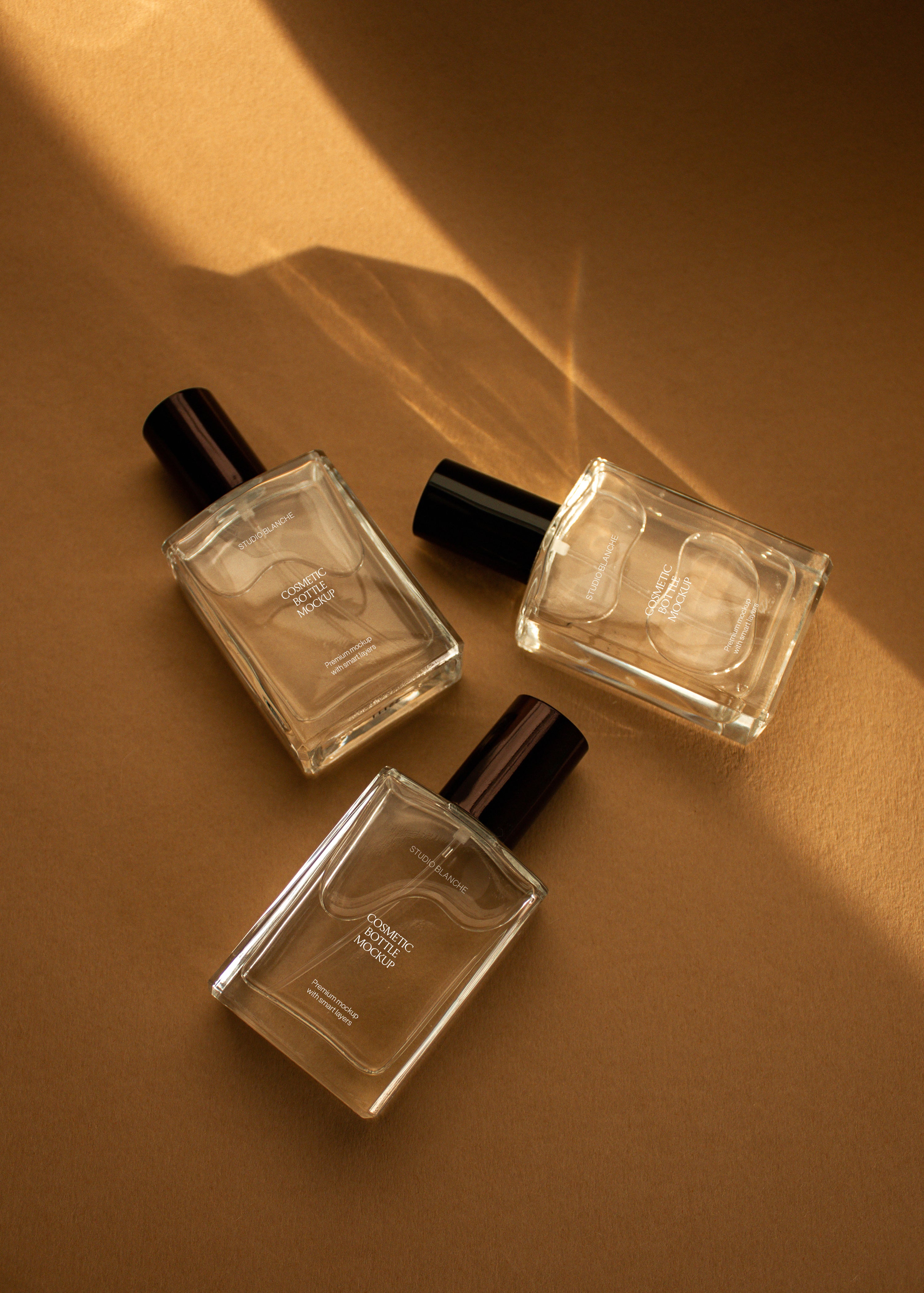 Trio of clear glass cosmetic bottle mockups with white and dark labels scattered on warm sandstone backdrop