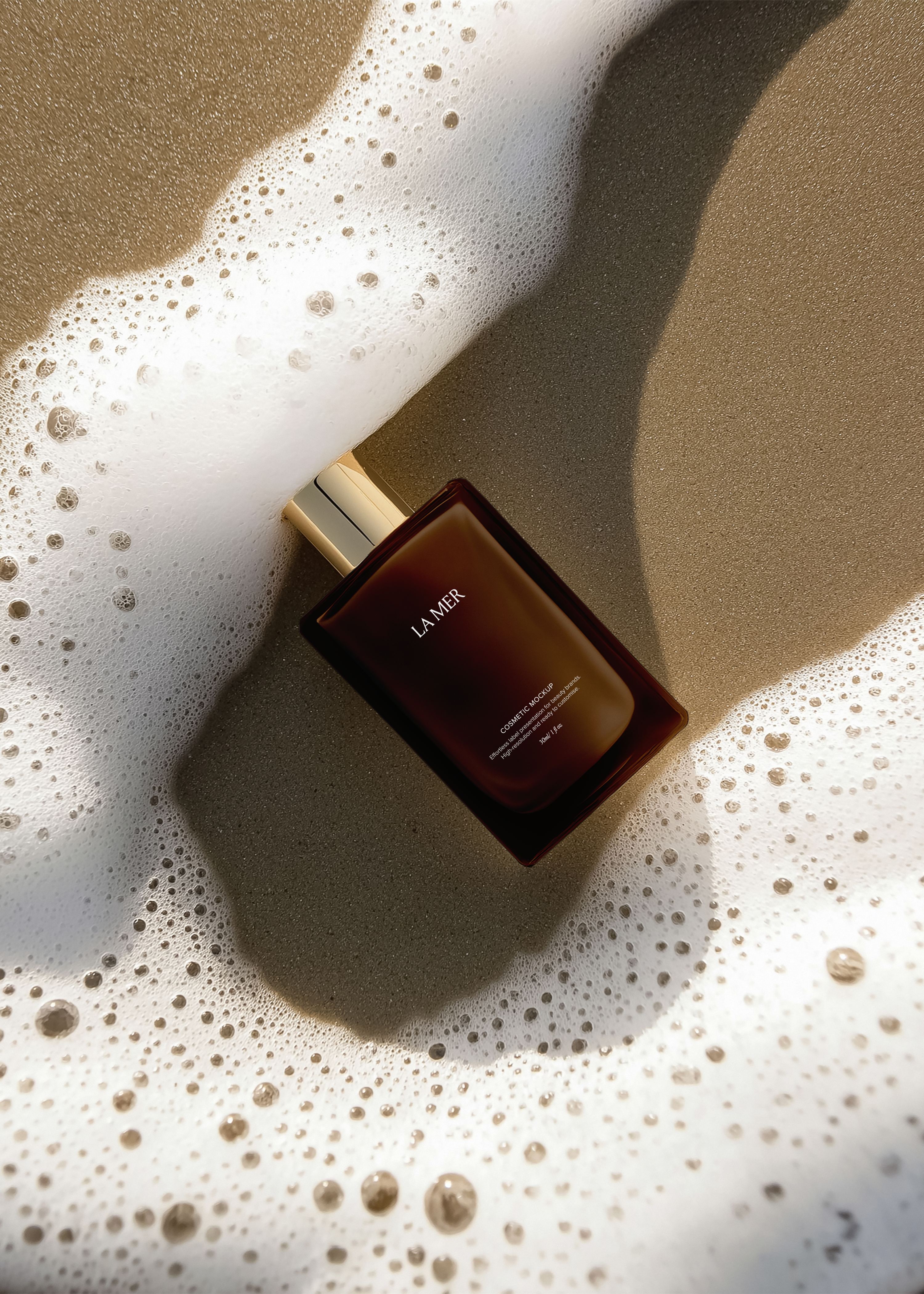 Dark amber cosmetic bottle with gold pump cap on wet sand, surrounded by white sea foam, flatlay overhead view
