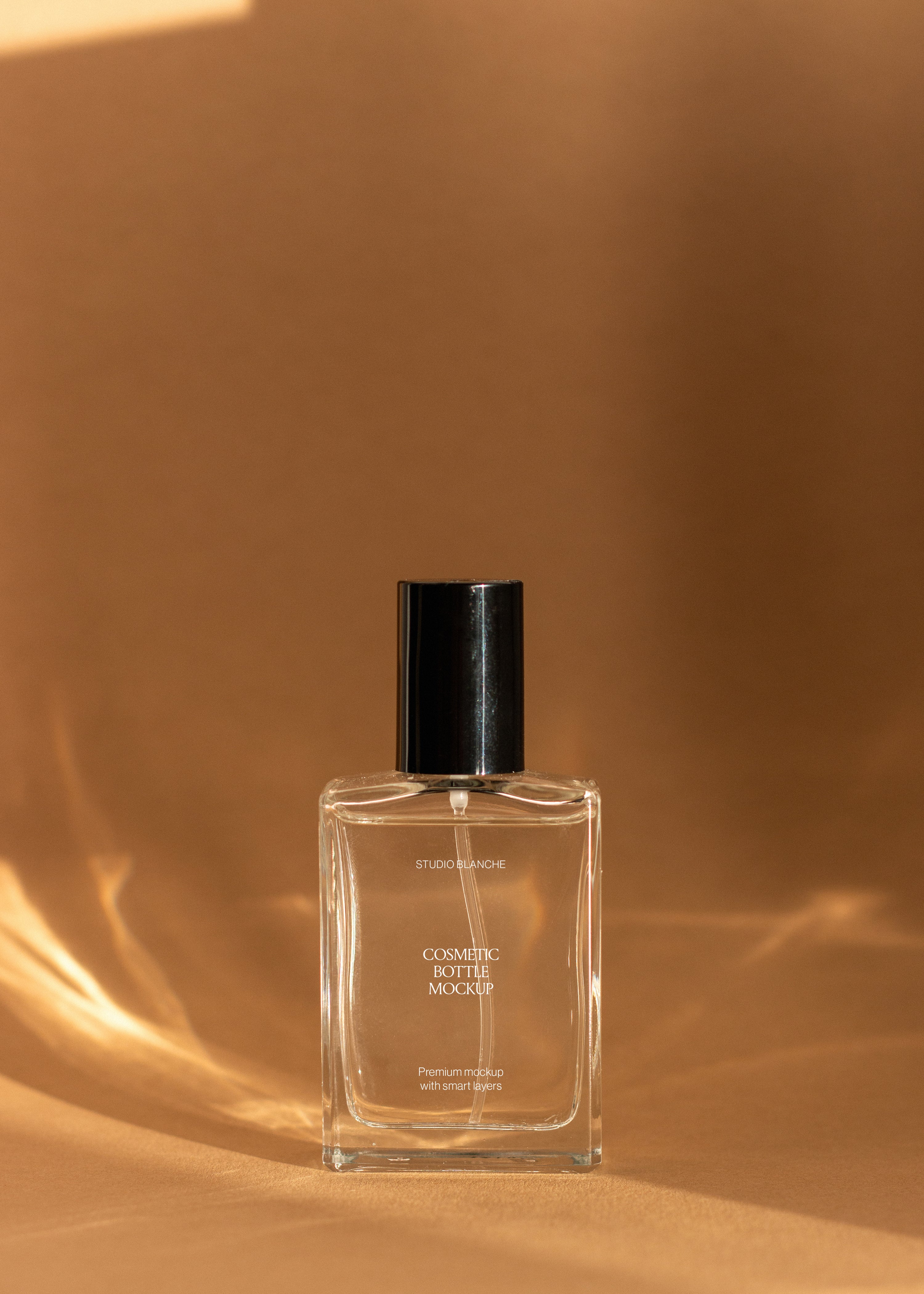 Clear glass cosmetic bottle mockup with black spray cap lit by soft warm light against draped sandstone fabric