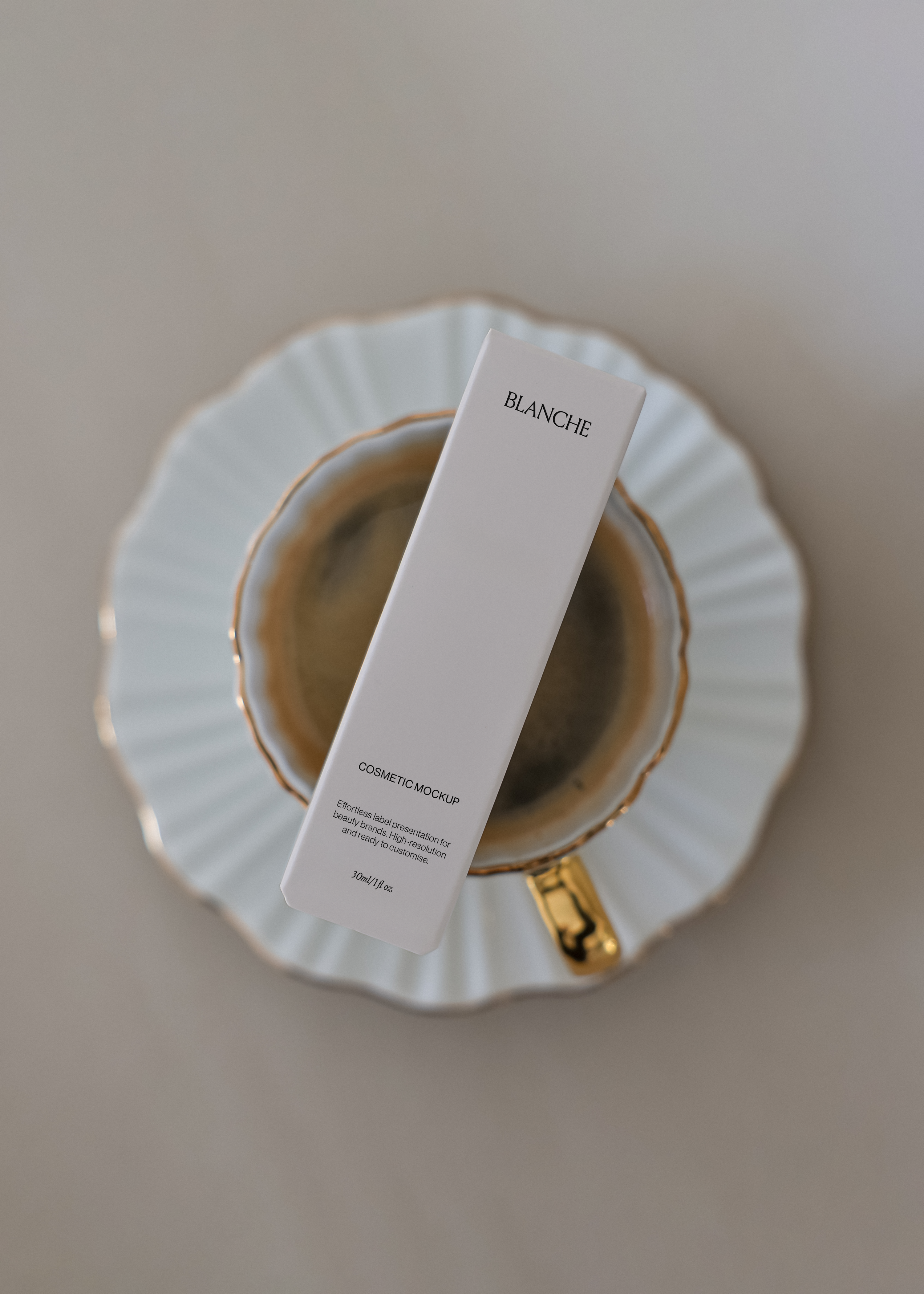 Tall white cosmetic box mockup laid diagonally across a gold-rimmed scalloped saucer and espresso cup on a warm beige surface