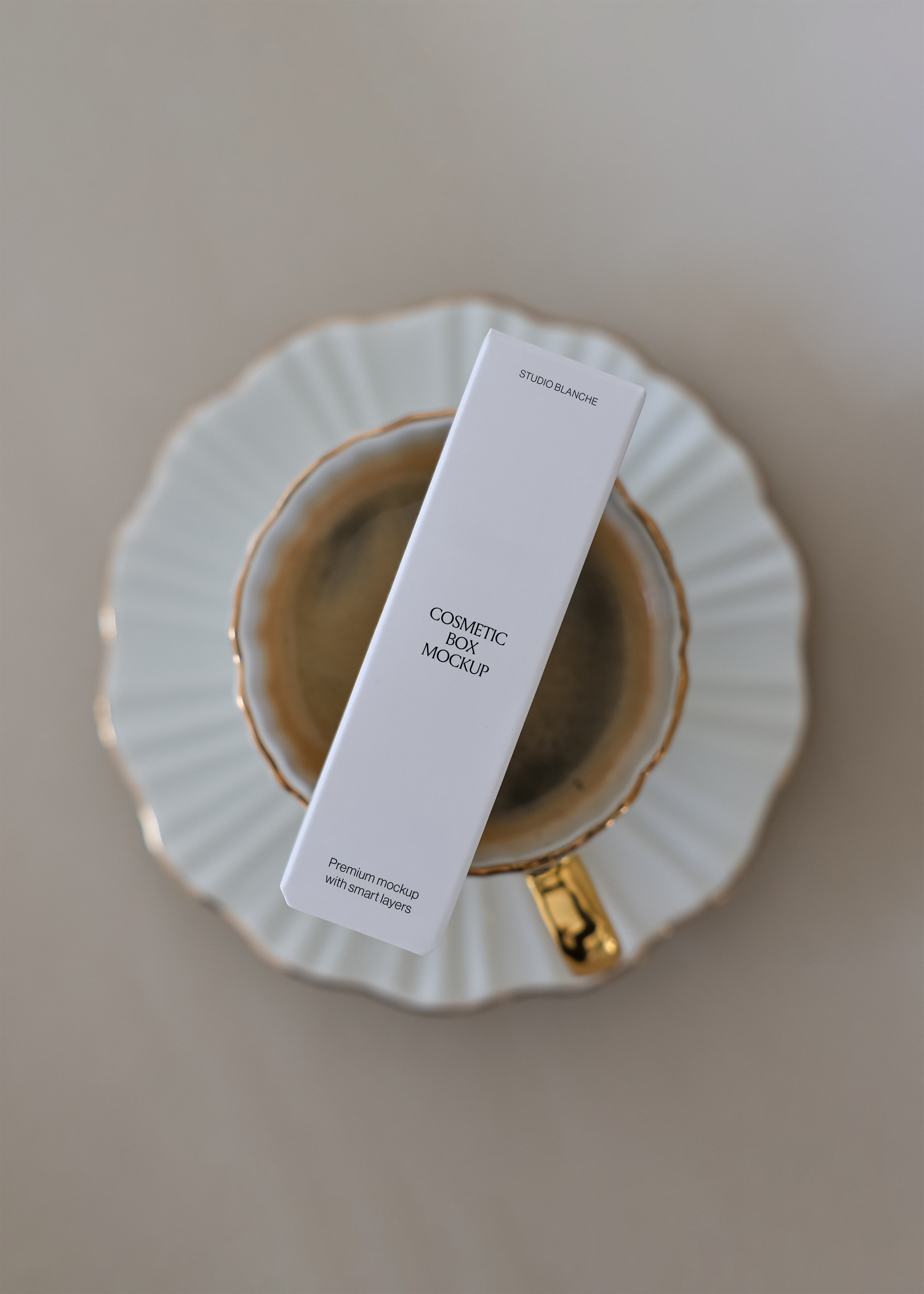 Close view of a minimal white cosmetic box mockup reading Studio Blanche resting on a fluted porcelain saucer with gold trim
