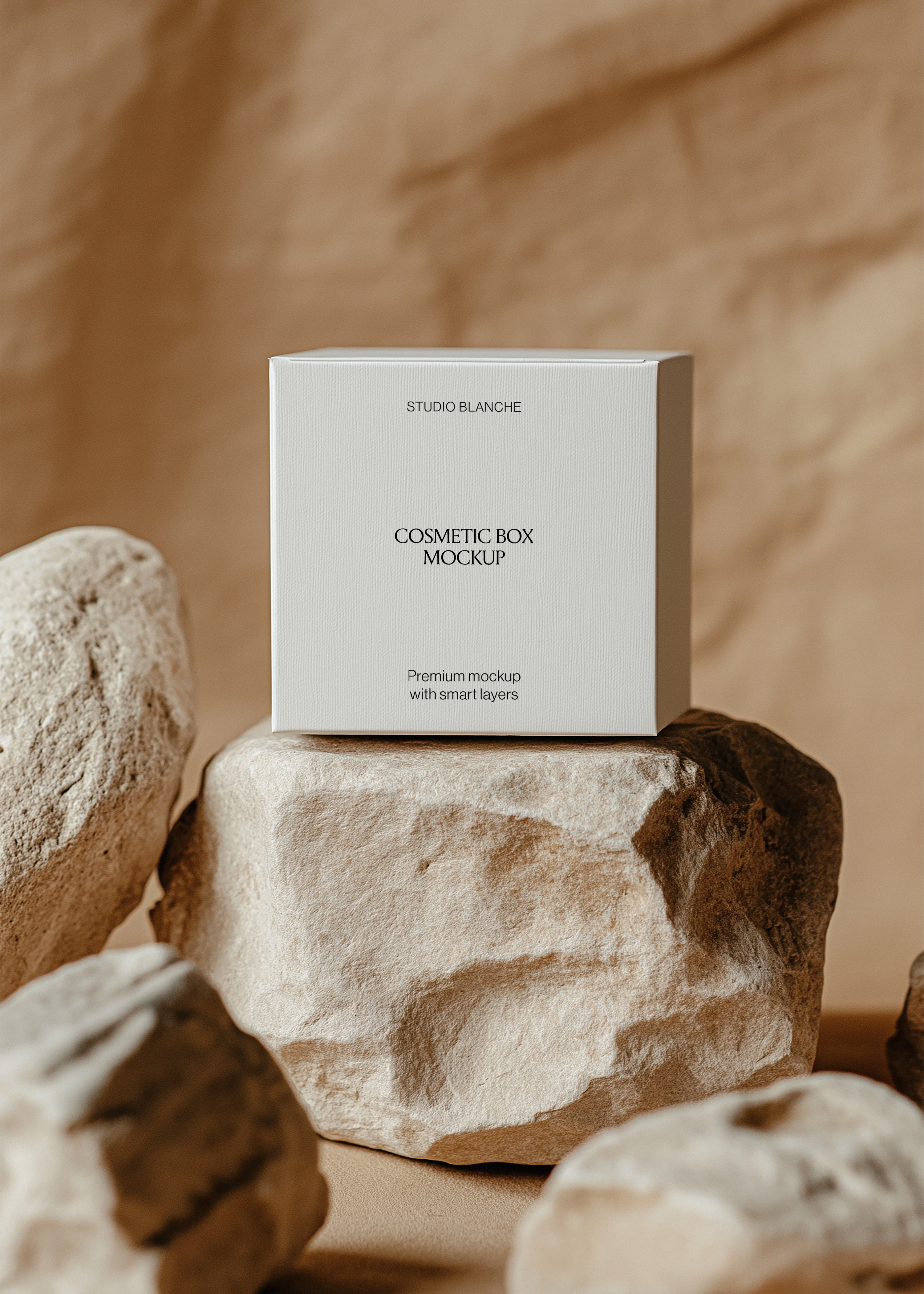 Front view of a white cosmetic box mockup reading Studio Blanche on a beige stone surrounded by rocks in warm neutral tones