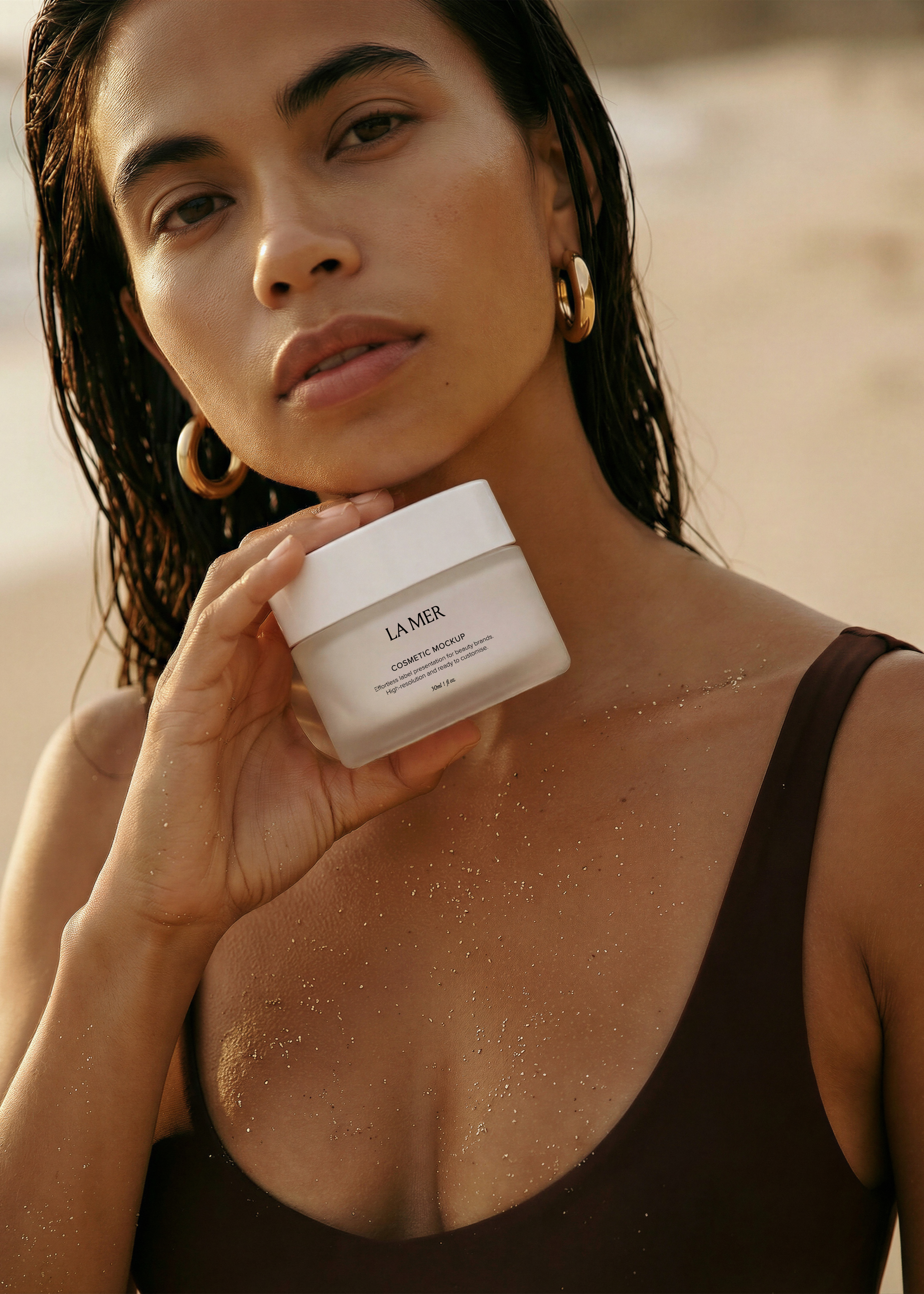 Cosmetic jar mockup held by model in brown swimsuit at the coast, frosted glass jar with label, warm golden-hour light