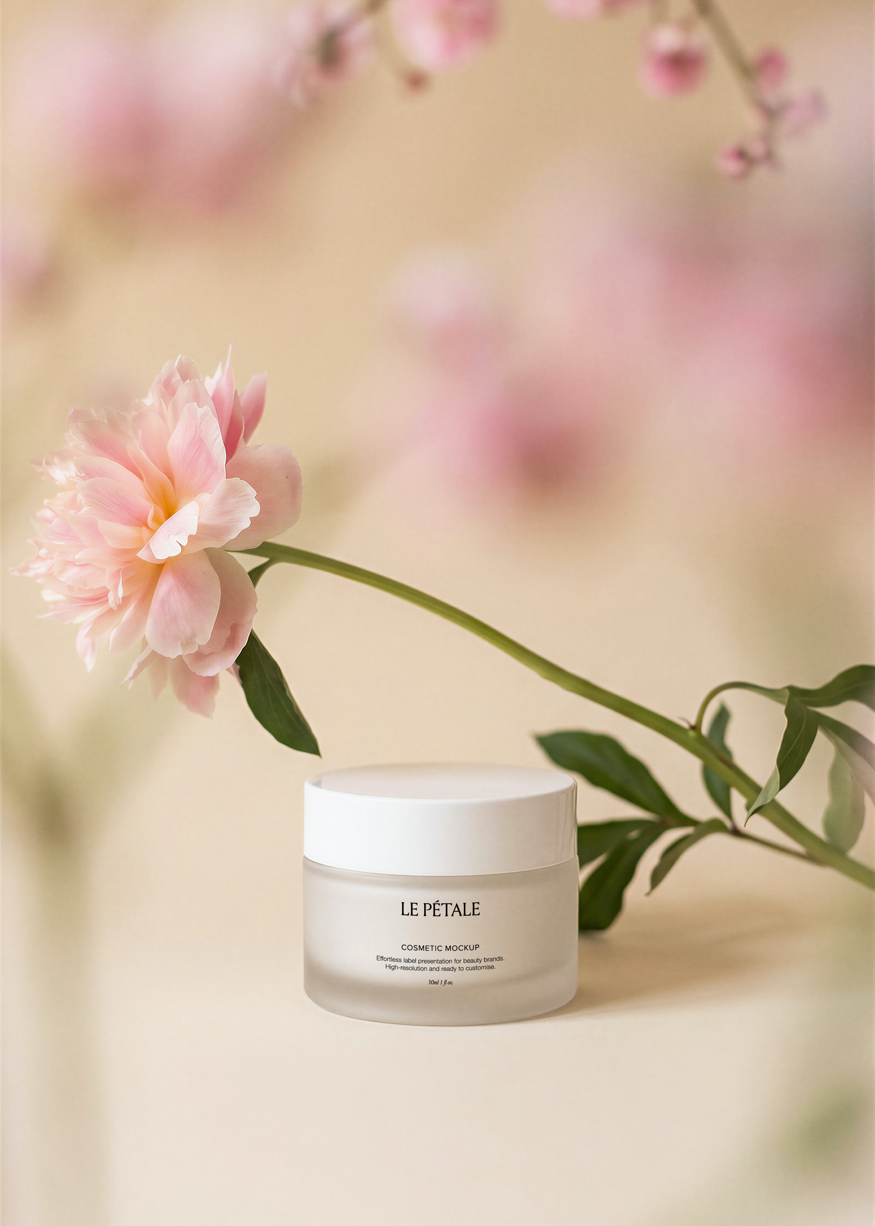 Frosted cosmetic jar mockup with white lid and label, pink peony stem, blurred blossom branches, warm sand backdrop