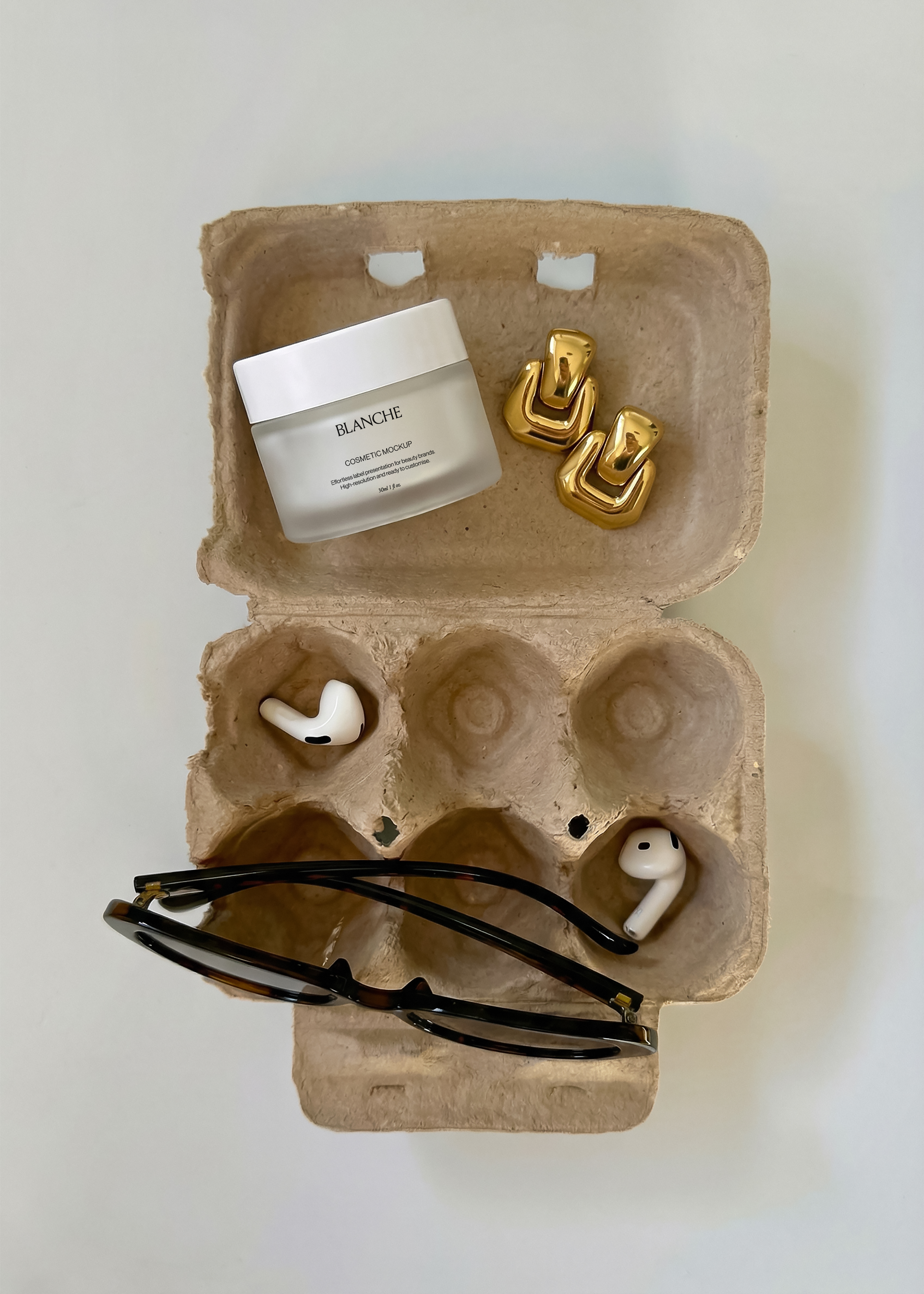 Frosted white cosmetic jar mockup in open egg carton with gold earrings, earbuds and tortoiseshell sunglasses