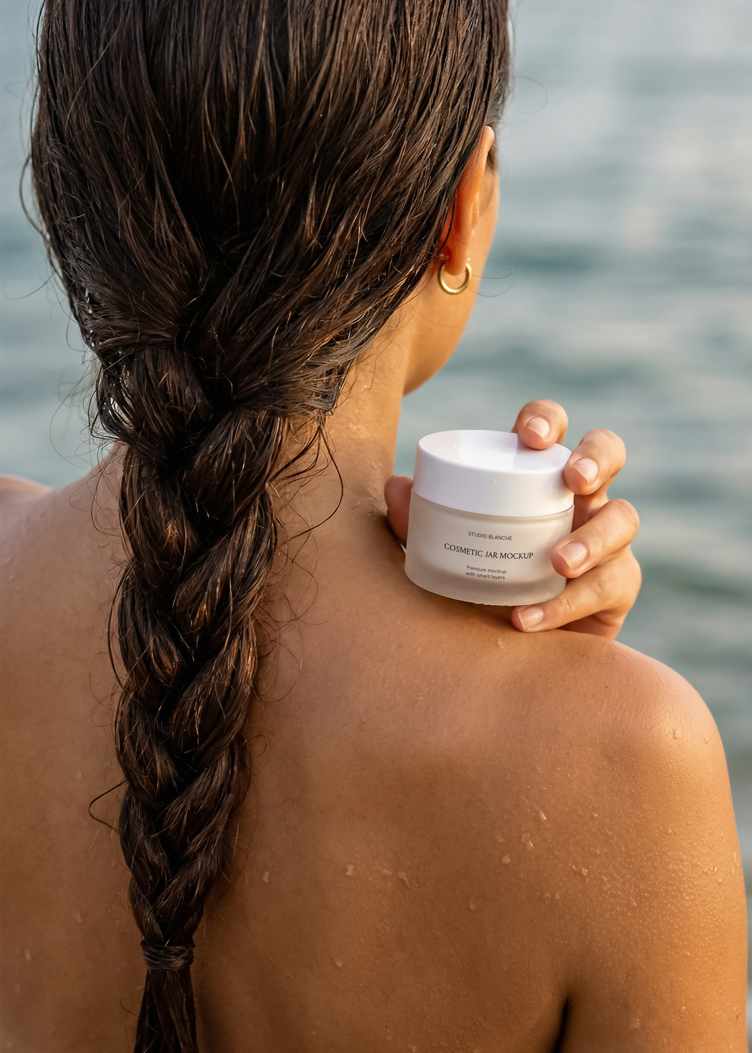 Frosted cosmetic jar mockup in coastal back portrait, wet braid, gold hoop earring, ocean backdrop, editorial styling