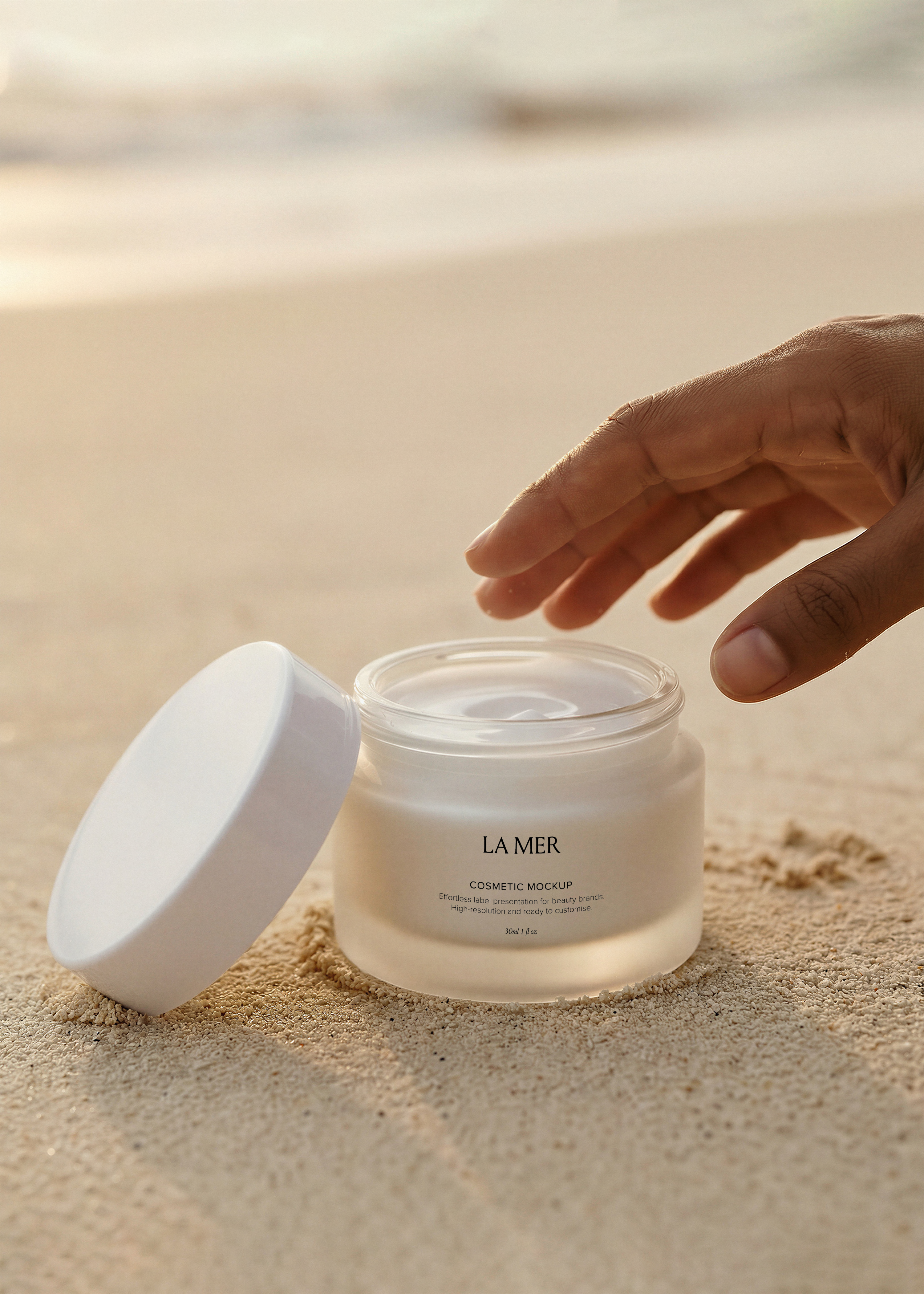 Open cosmetic jar mockup on soft sand with lid resting beside, hand reaching toward cream, golden-hour coastal light