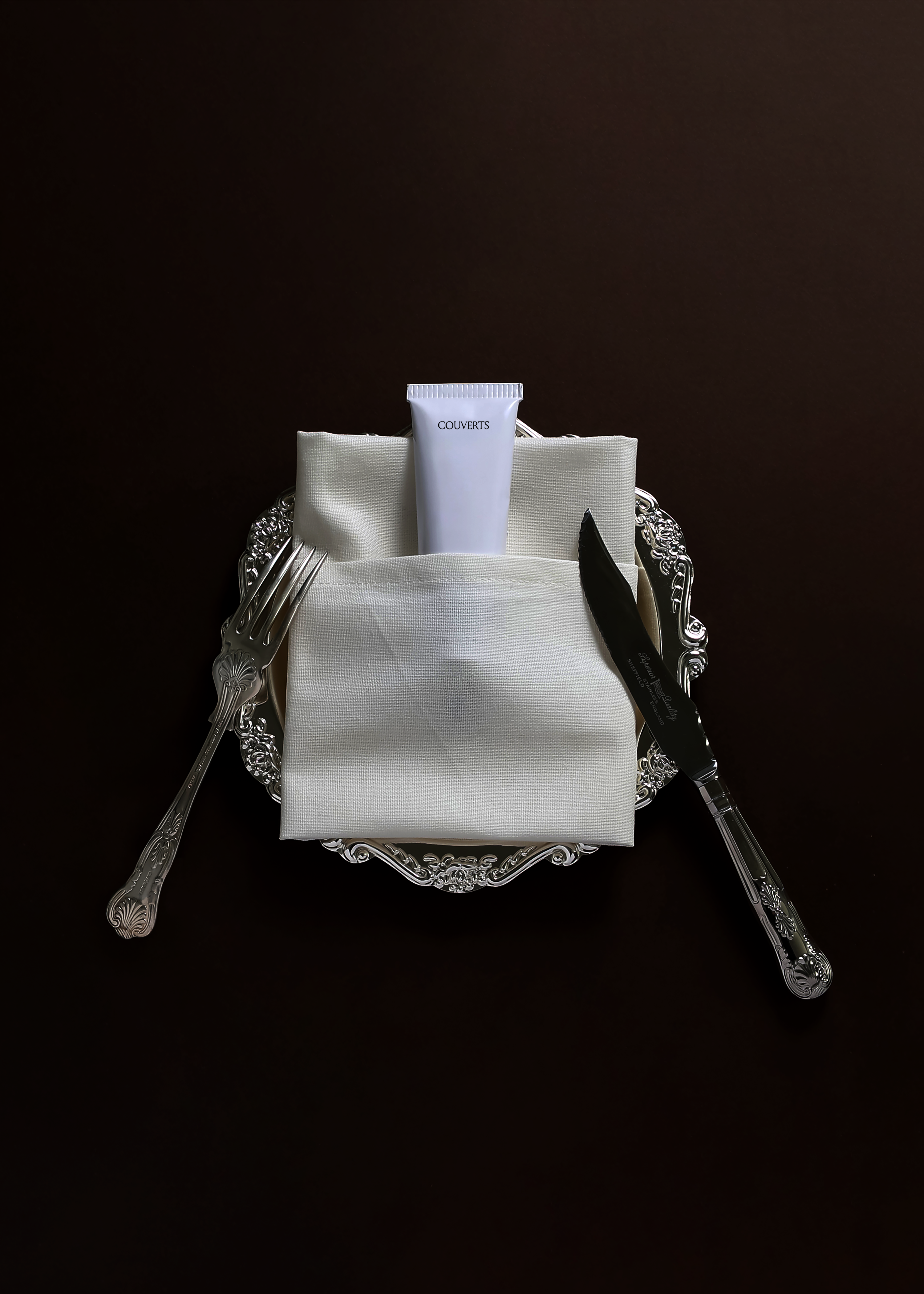 Cosmetic tube mockup tucked into a folded linen napkin on an ornate silver plate with fork and knife, dark brown backdrop