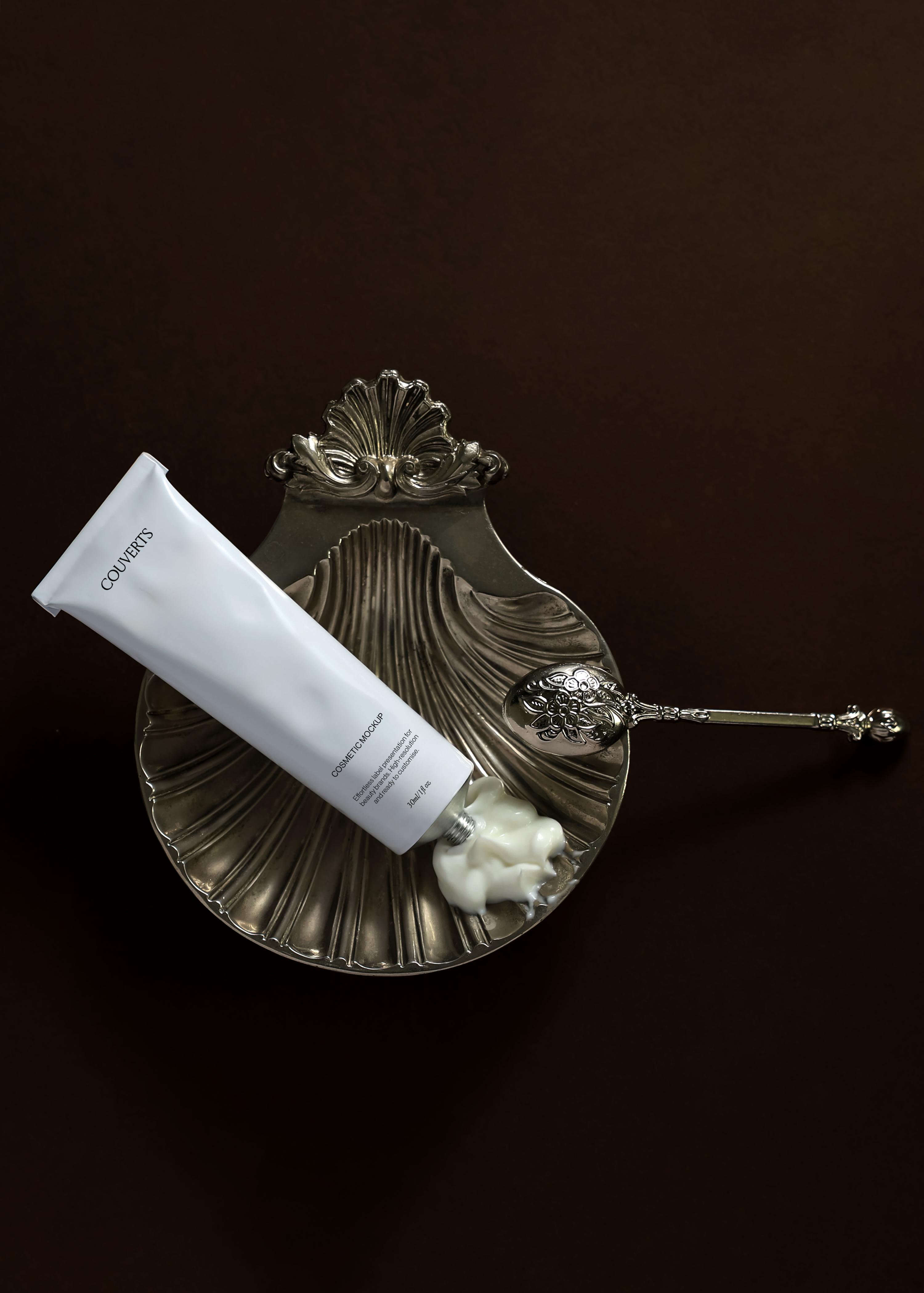 Cosmetic tube mockup resting on an ornate silver shell dish with a floral spoon and cream swirl, dark backdrop