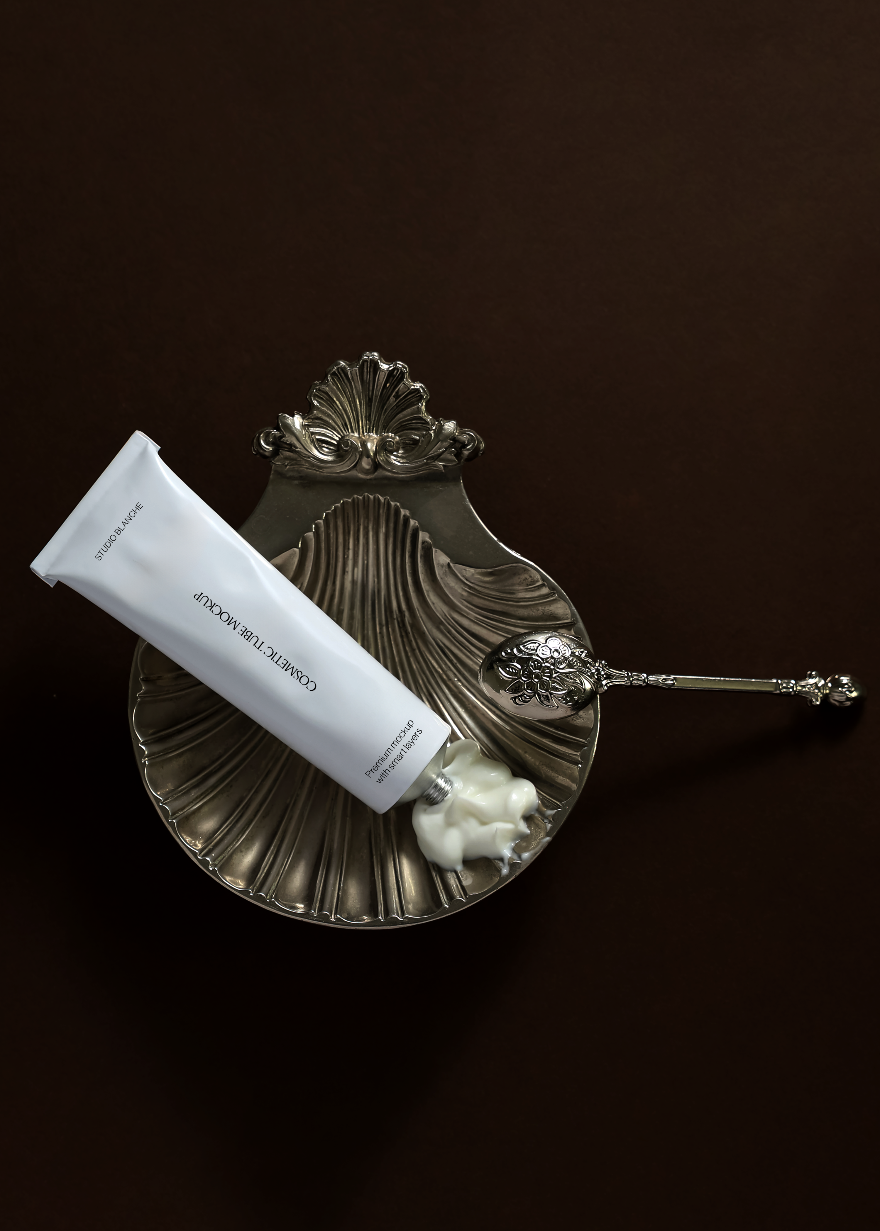 White cosmetic tube mockup with cream dispensed onto an antique silver shell dish, engraved spoon, dark brown background