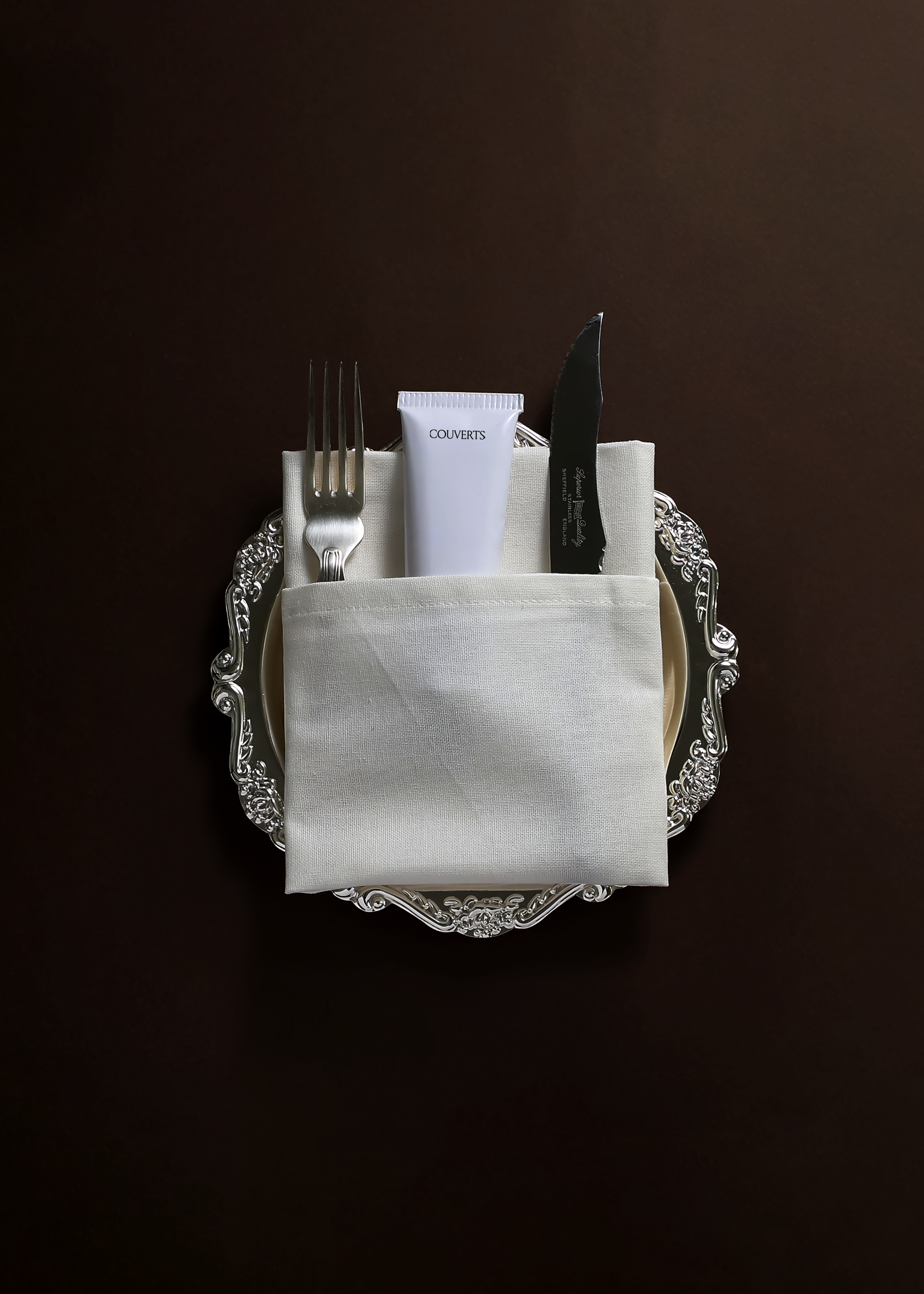 Cosmetic tube mockup in a folded linen napkin with silver fork and knife on an ornate plate, dark brown backdrop