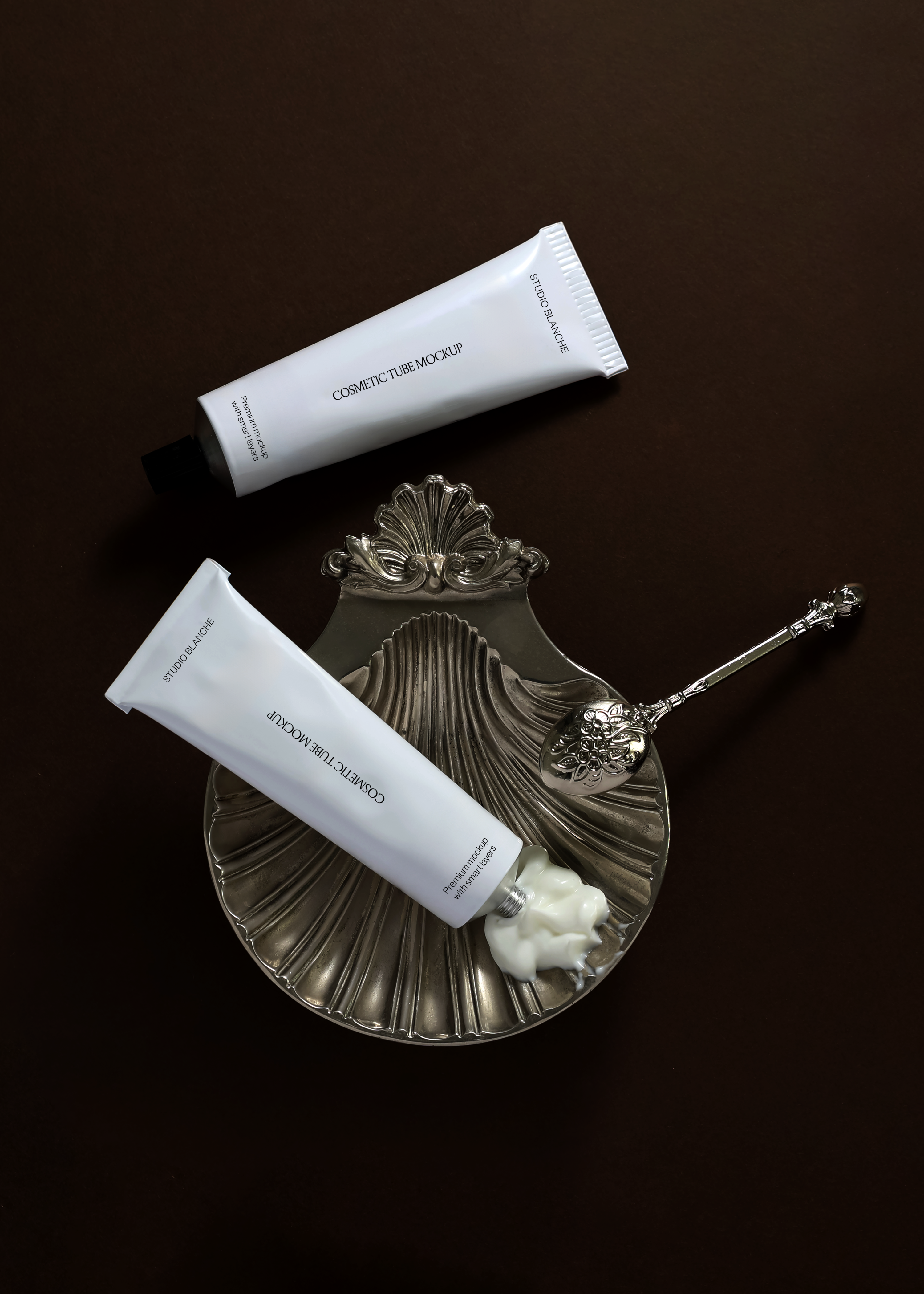 White cosmetic tube duo mockup, one resting on an engraved silver shell dish with dispensed cream, top-down view