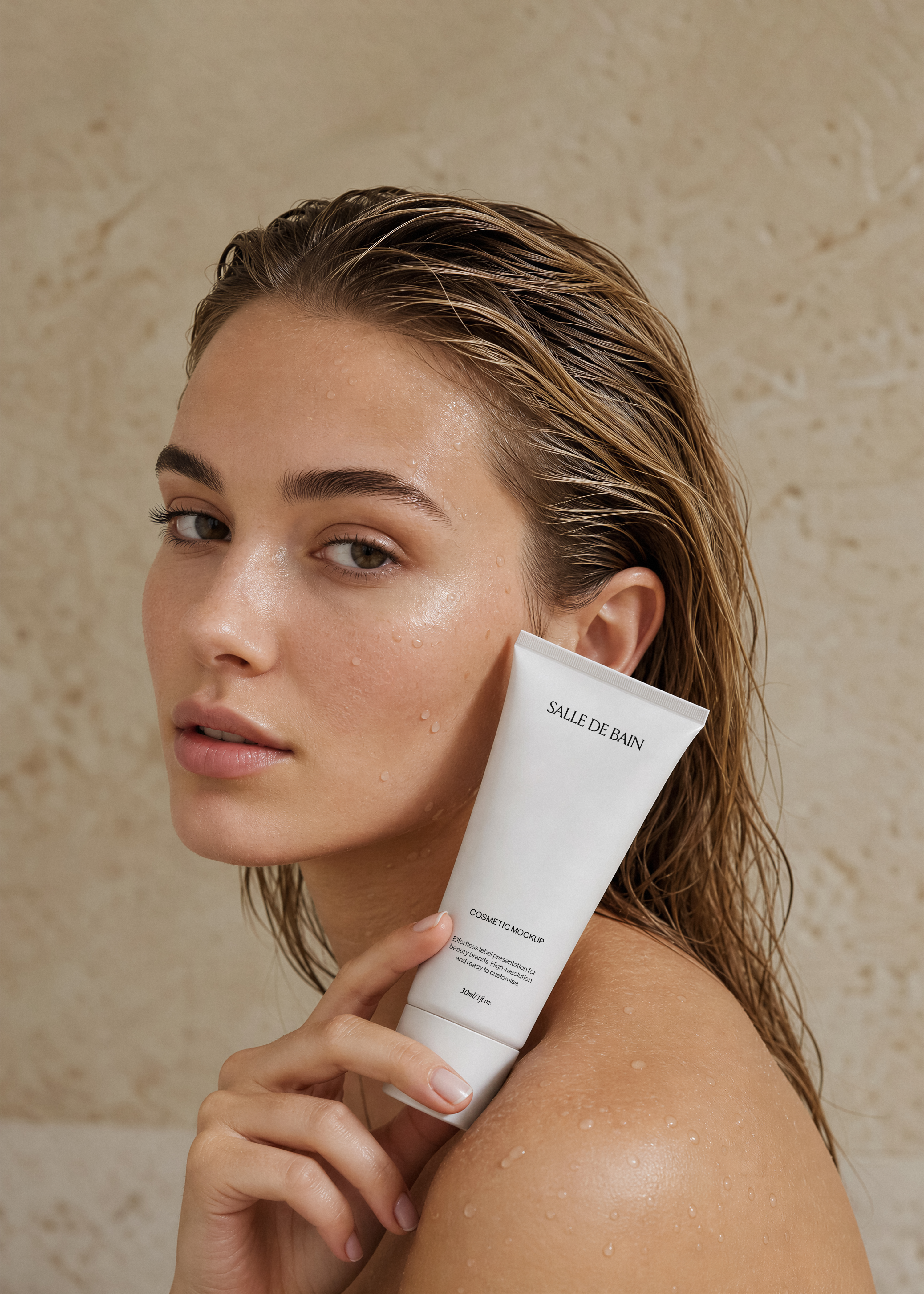 Cosmetic tube mockup held against the cheek of a model with wet hair and dewy skin, warm sandstone wall behind