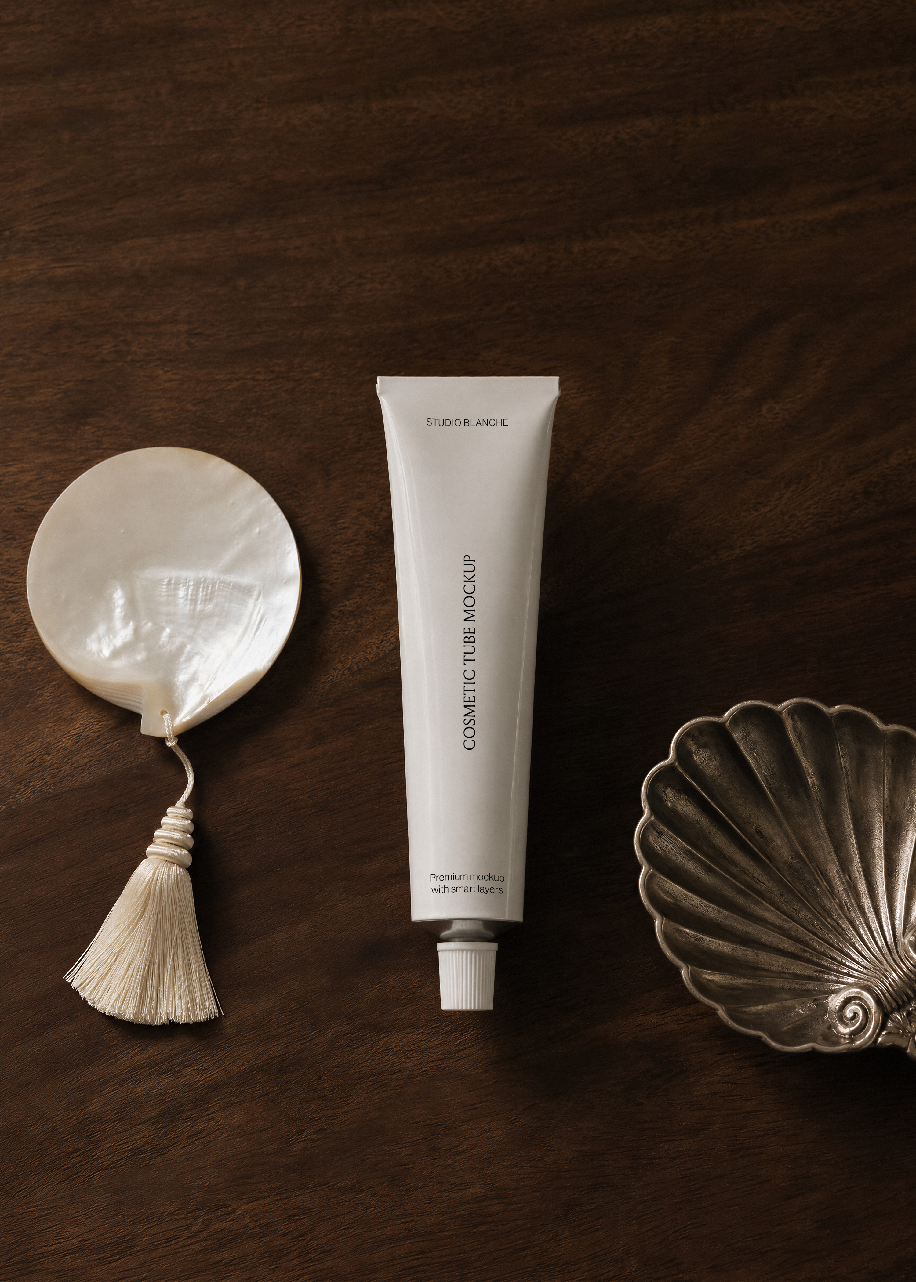 White cosmetic tube mockup on dark walnut surface beside mother-of-pearl disc with cream tassel and antique silver scallop dish