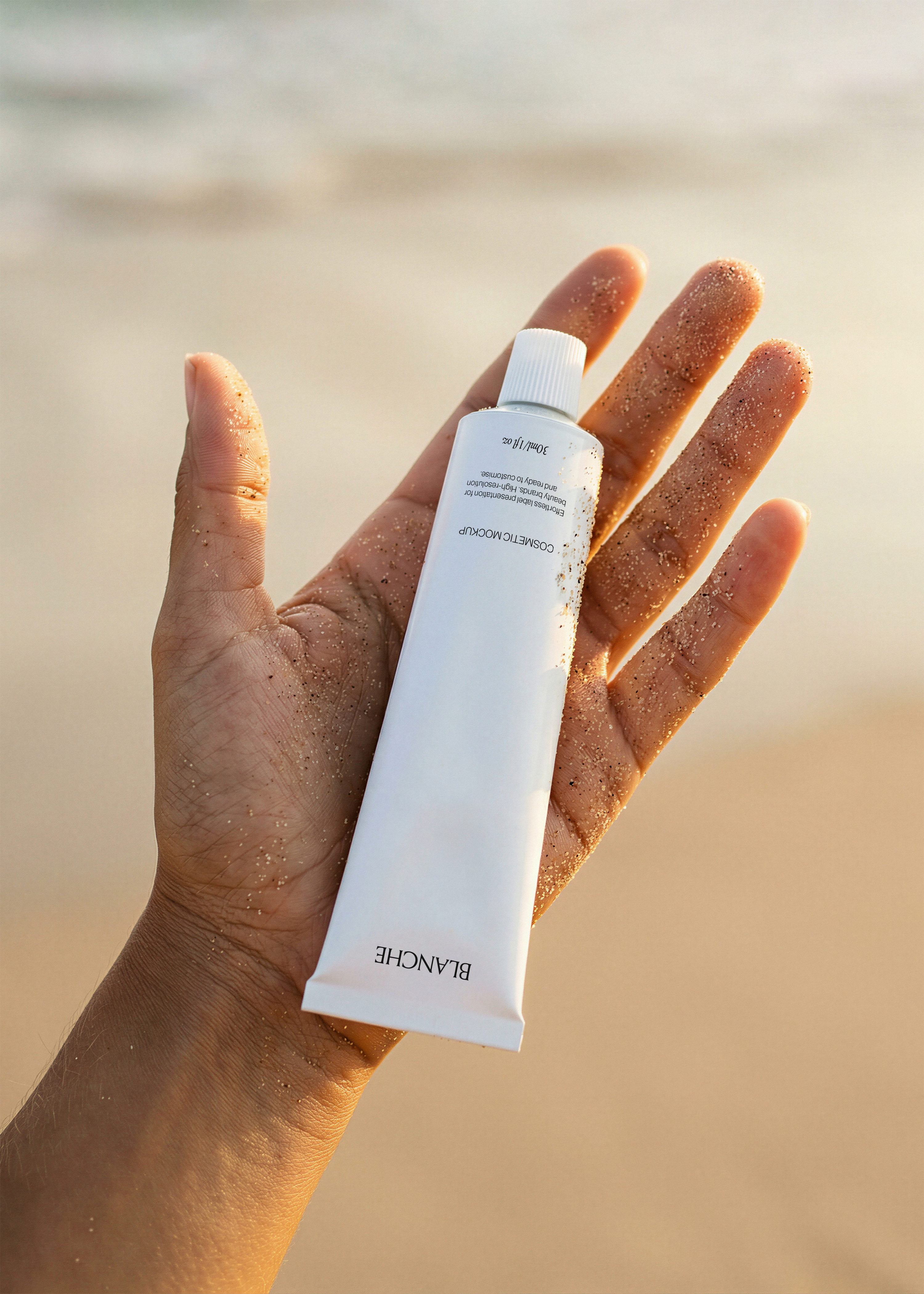 White cosmetic tube mockup held in a sand-covered hand against a blurred beach shoreline at golden hour