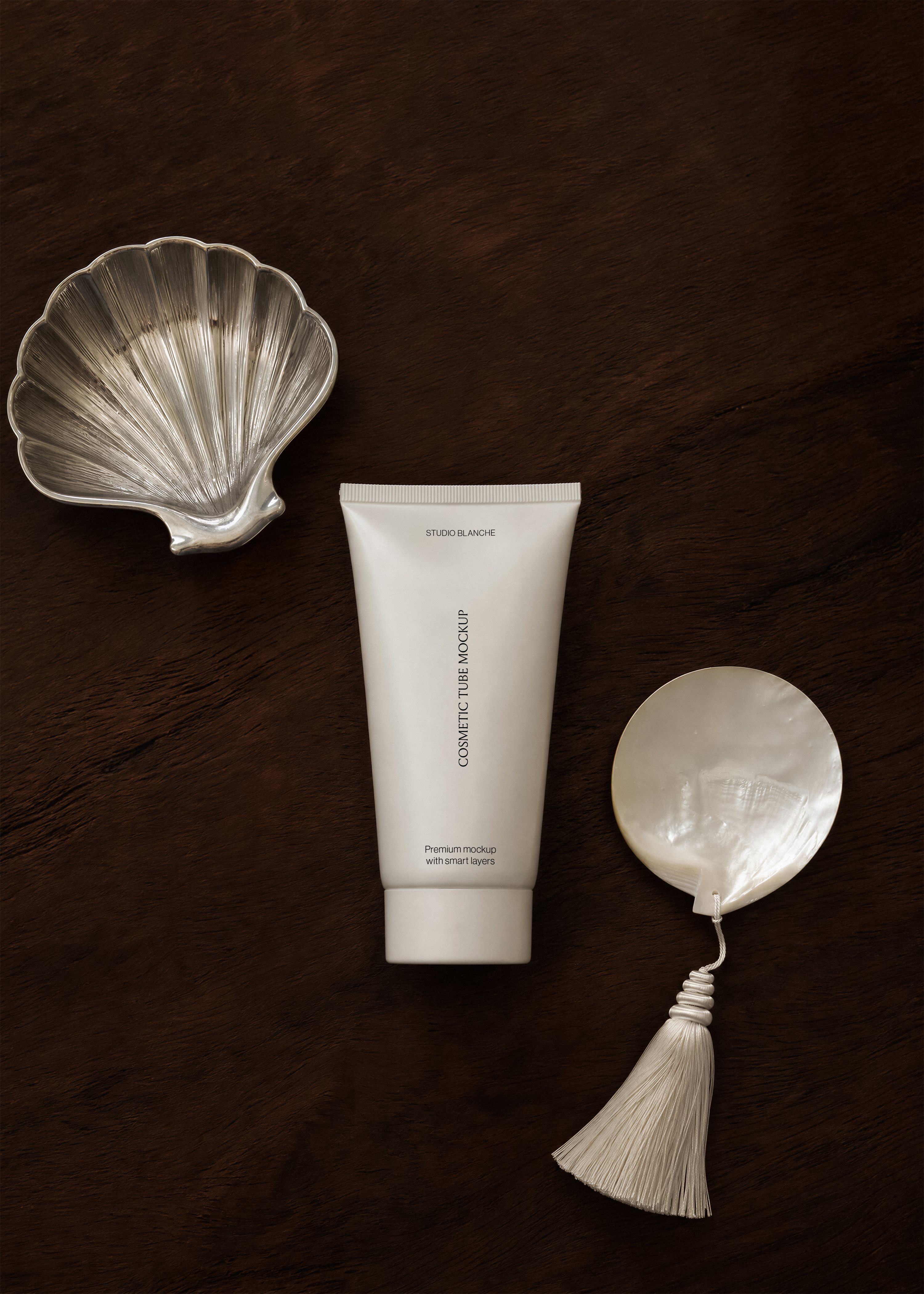 White cosmetic tube mockup on dark walnut surface beside antique silver scallop dish and mother-of-pearl disc with cream tassel