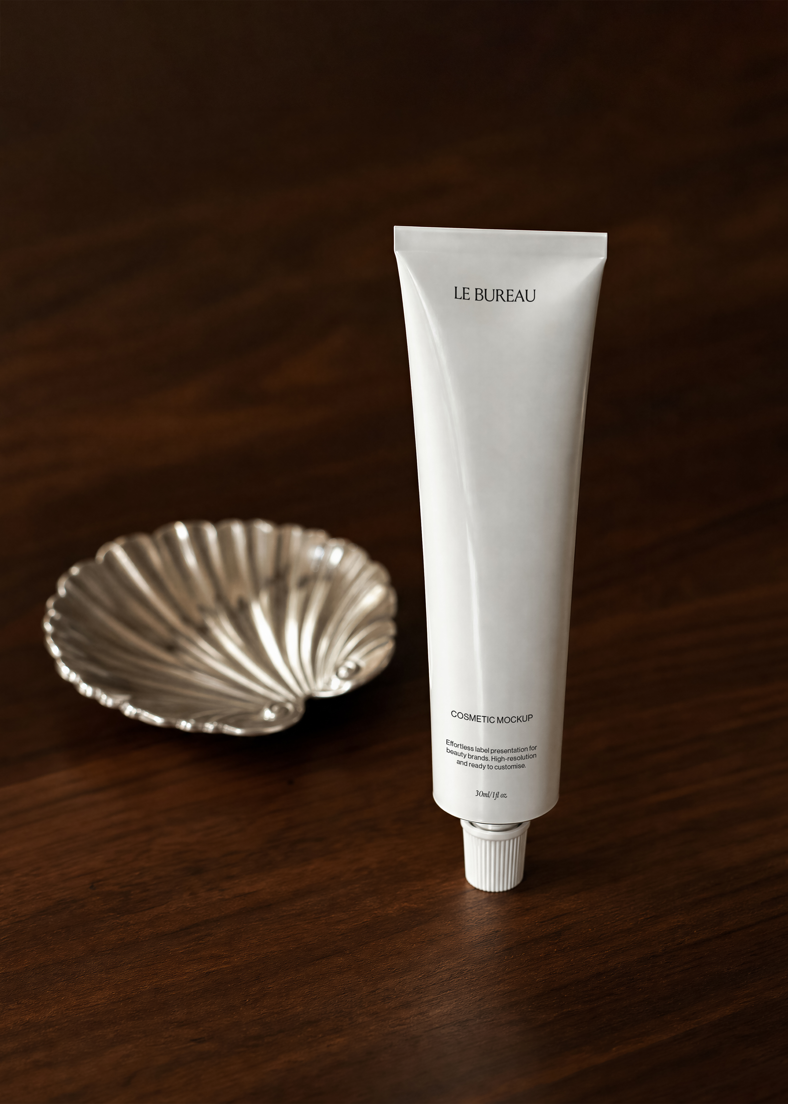 Cosmetic tube mockup displaying Le Bureau branding upright on dark walnut surface, antique silver shell dish beside it