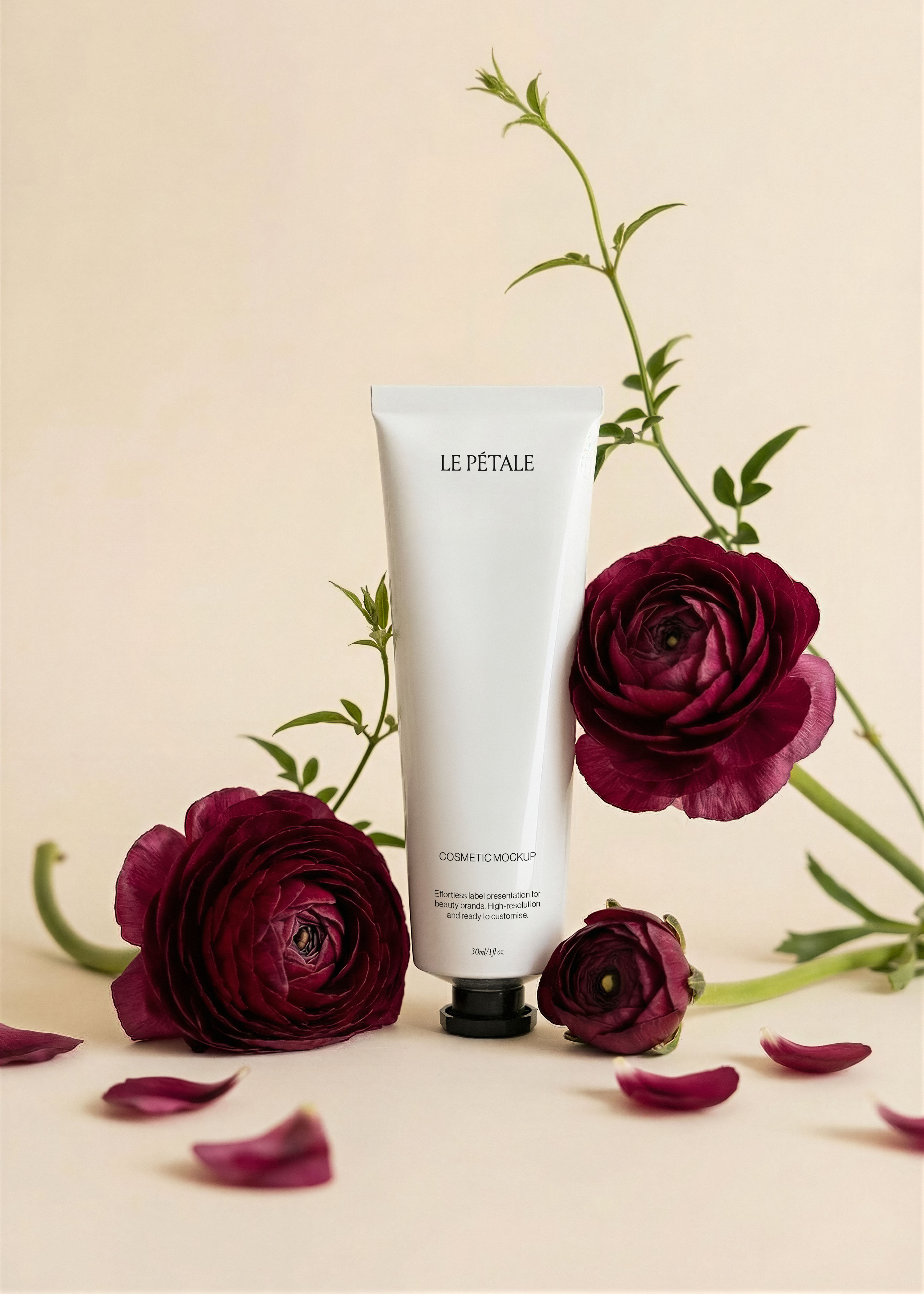 Cosmetic tube mockup with label, burgundy ranunculus blooms flanking, scattered petals, green stem, warm sand backdrop