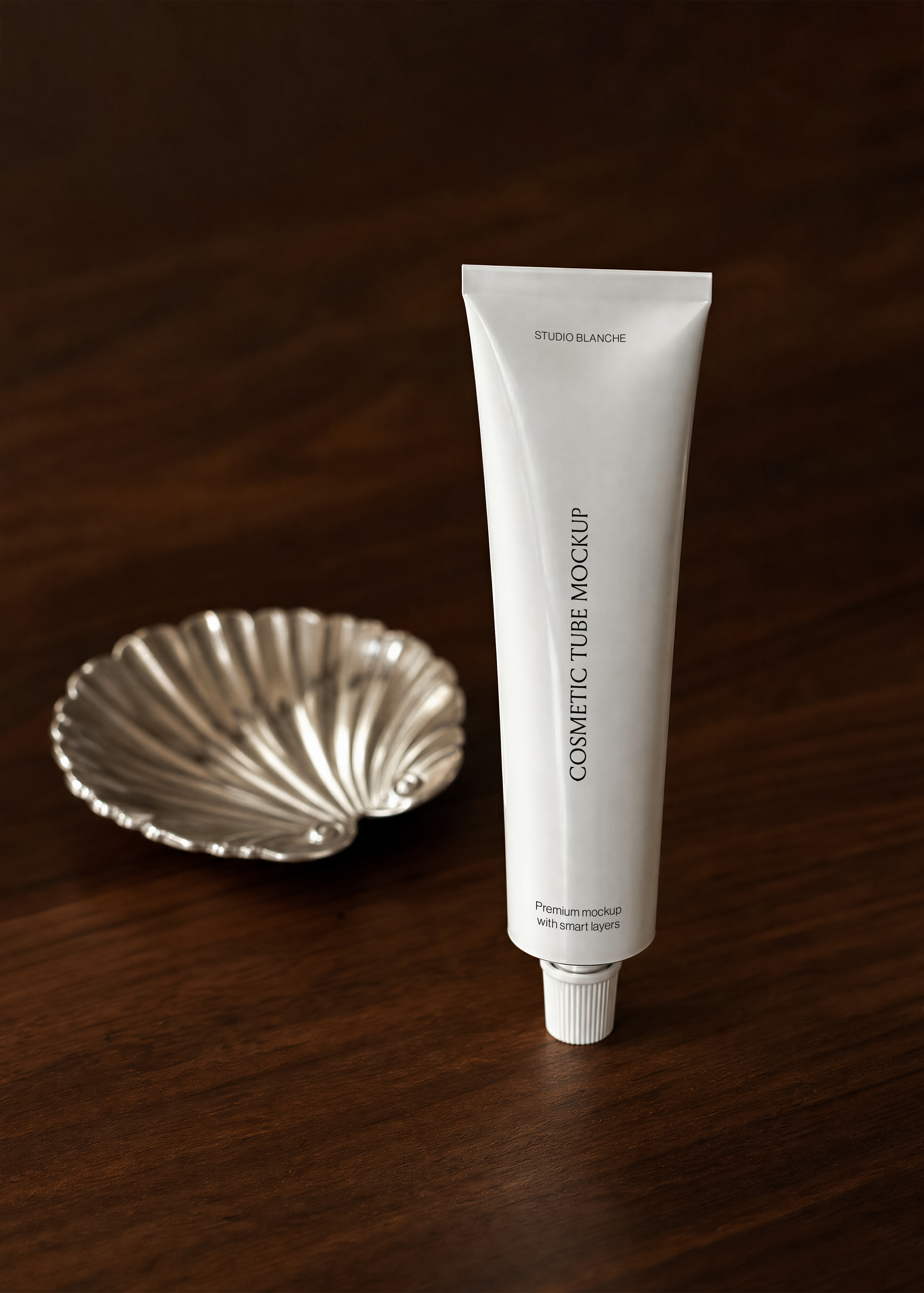 White cosmetic tube mockup standing upright on dark espresso walnut surface, antique silver scallop dish set back beside it