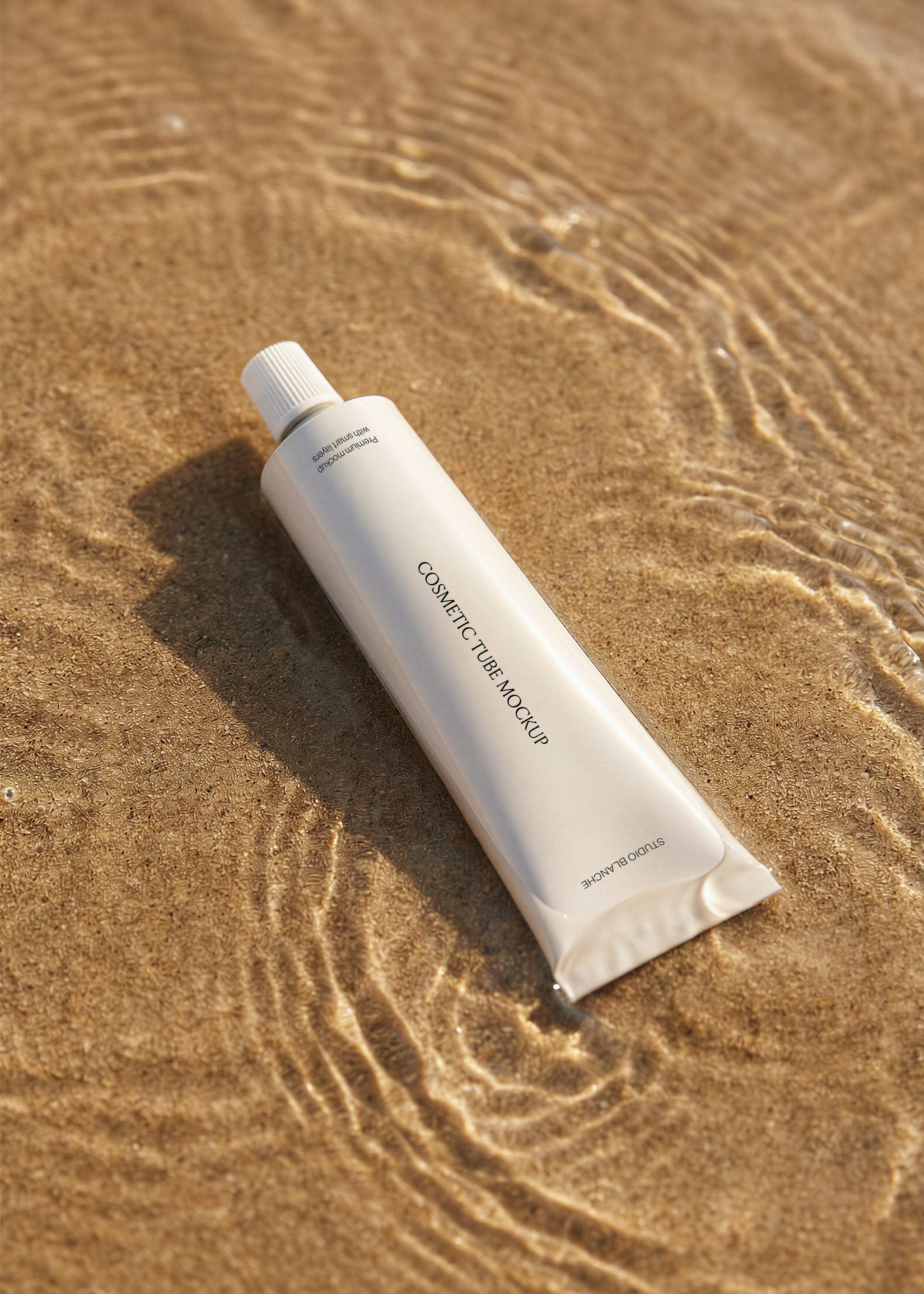 White cosmetic tube mockup resting on sun-lit rippled wet sand with a long cast shadow on a beach