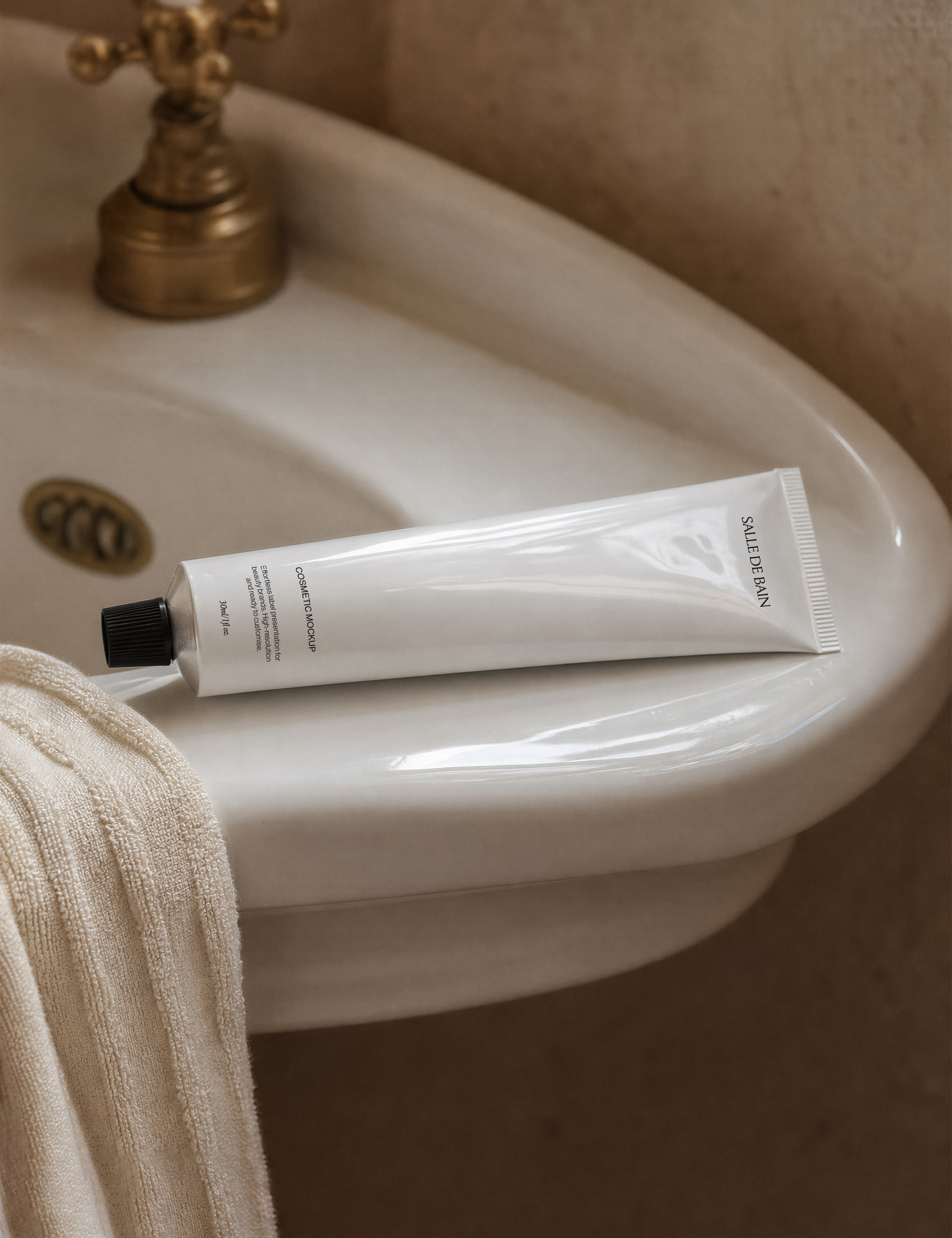 White cosmetic tube mockup with black cap on porcelain pedestal sink edge, brass tap and draped cream towel, warm light