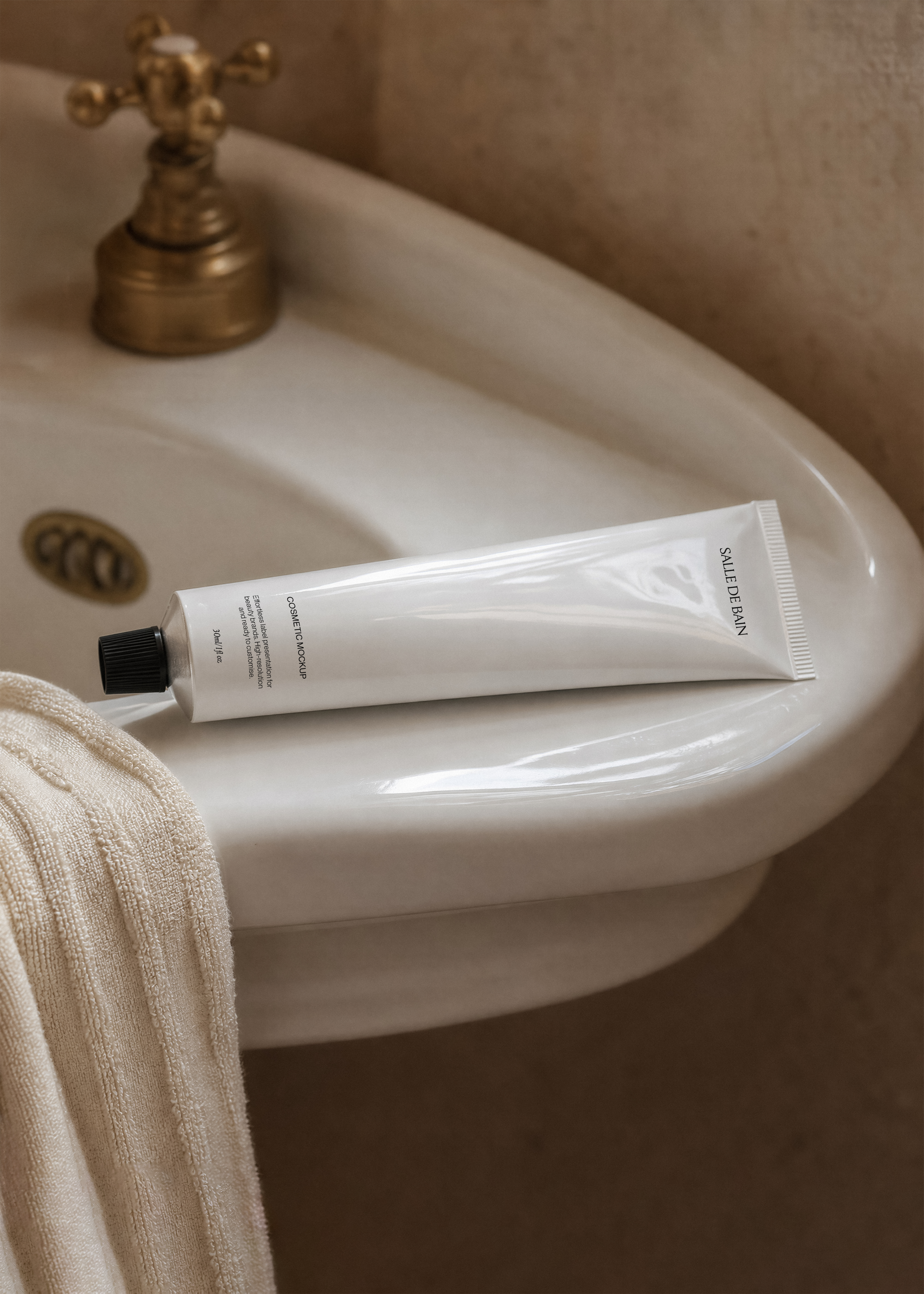 White cosmetic tube mockup with black cap on porcelain pedestal sink edge, brass tap and draped cream towel, warm light